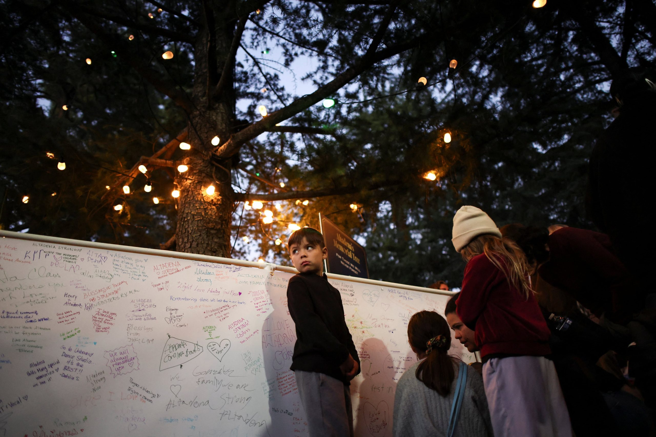 Un niño observa mientras la gente deja mensajes en una pancarta blanca que rodea un árbol conmemorativo en memoria de las víctimas del incendio de Eaton en el evento anual de iluminación de Christmas Tree Lane en Altadena, California.