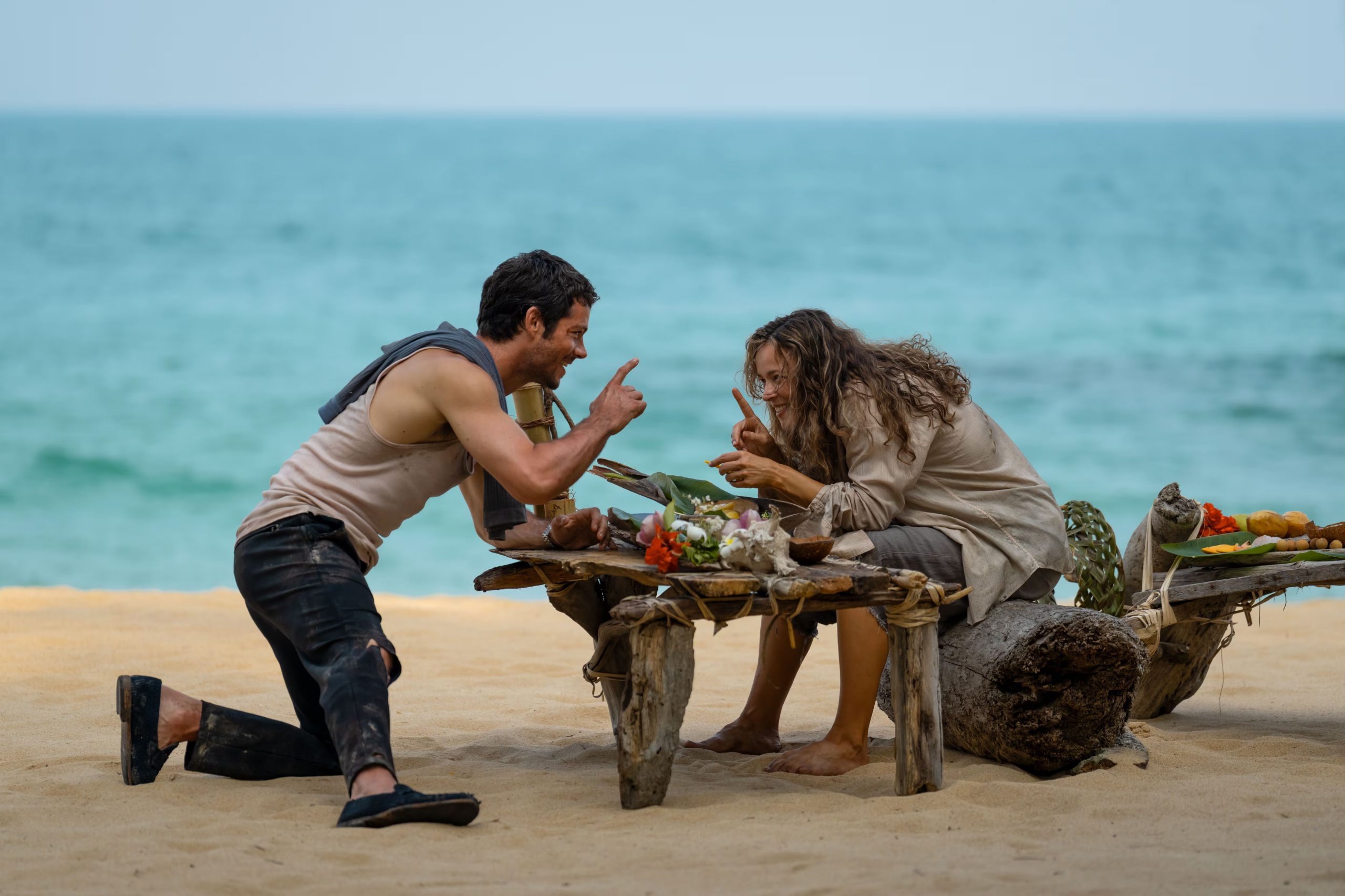 Un hombre y una mujer, aparentemente náufragos, comparten un momento intenso de comunicación sobre una mesa improvisada en la orilla de una isla remota, quizás buscando una solución o esperando AYUDA (Disney)