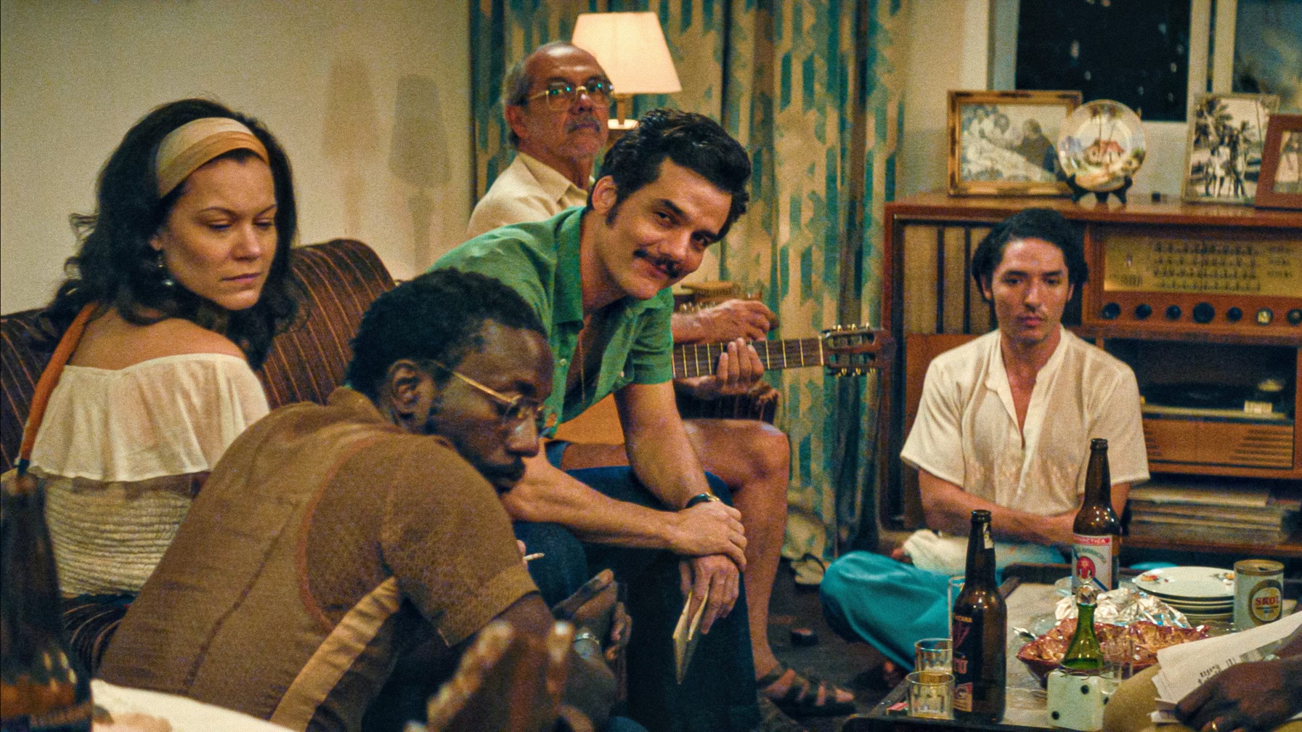 Wagner Moura, Emilie Lesclaux y otros miembros del elenco disfrutan de una reunión social en un decorado que evoca los años setenta, compartiendo música y compañía. (MUBI)