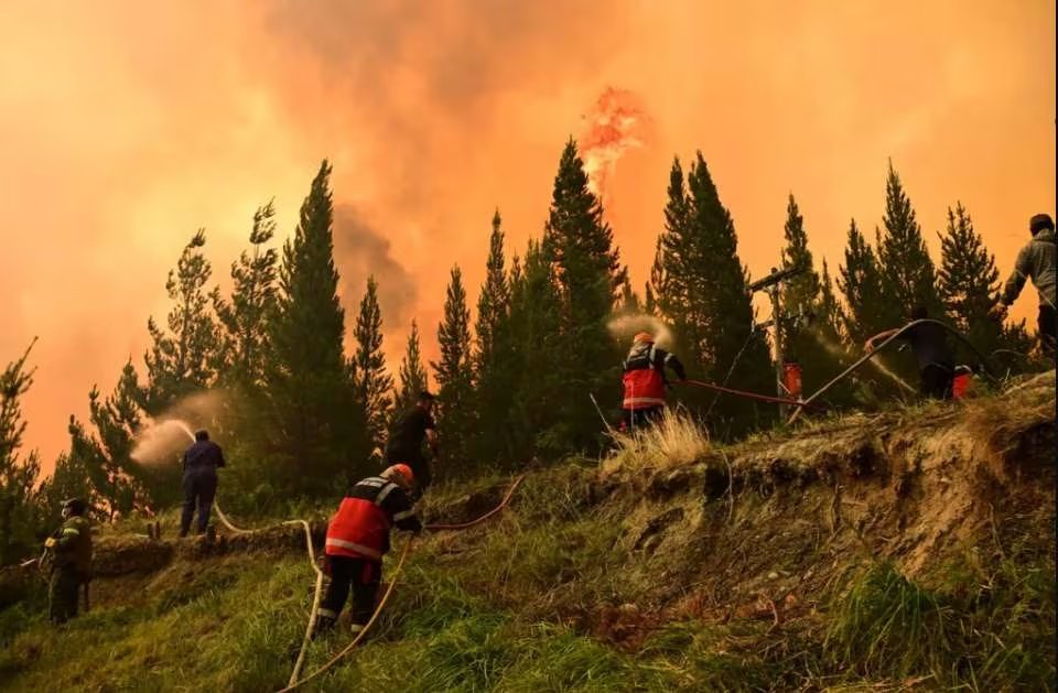 Un gran incendio forestal en Chubut emite intensas llamas y humo naranja, mientras vehículos de emergencia se congregan en una carretera cercana listos para actuar (Imagen Ilustrativa Infobae)