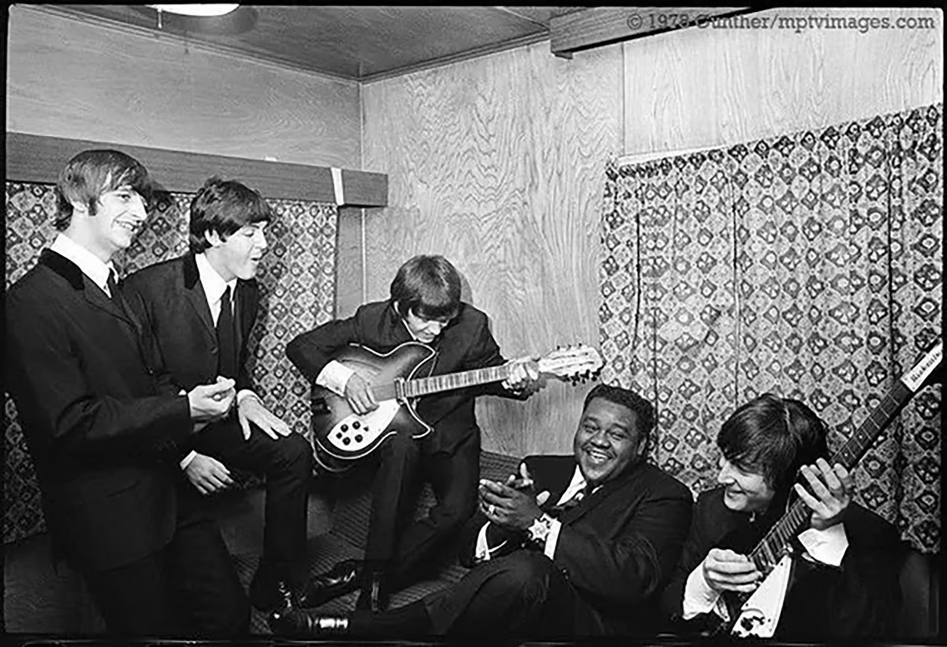 Fats Domino sonríe mientras observa a The Beatles tocar la guitarra en un animado encuentro tras bambalinas, un momento histórico para la música