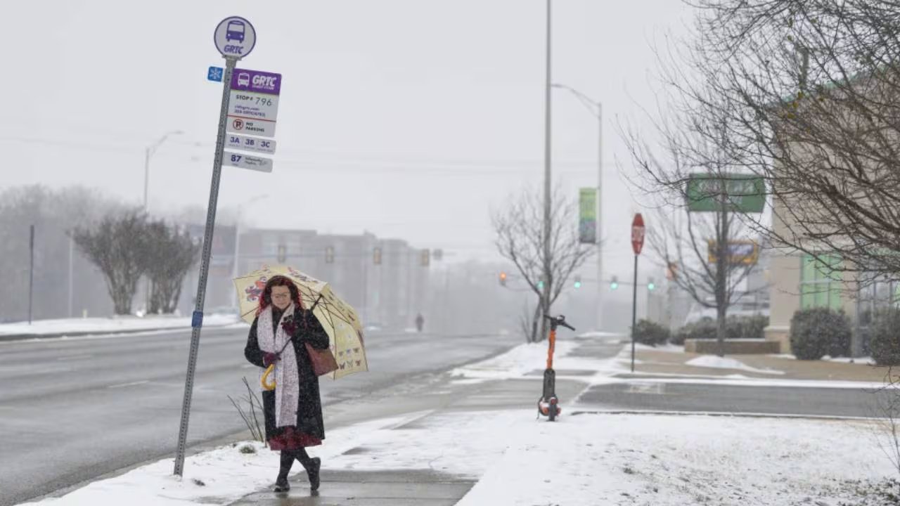 Margo Wagner camina por una acera cubierta de nieve con un paraguas durante una ventisca invernal, esperando en una parada de autobús.