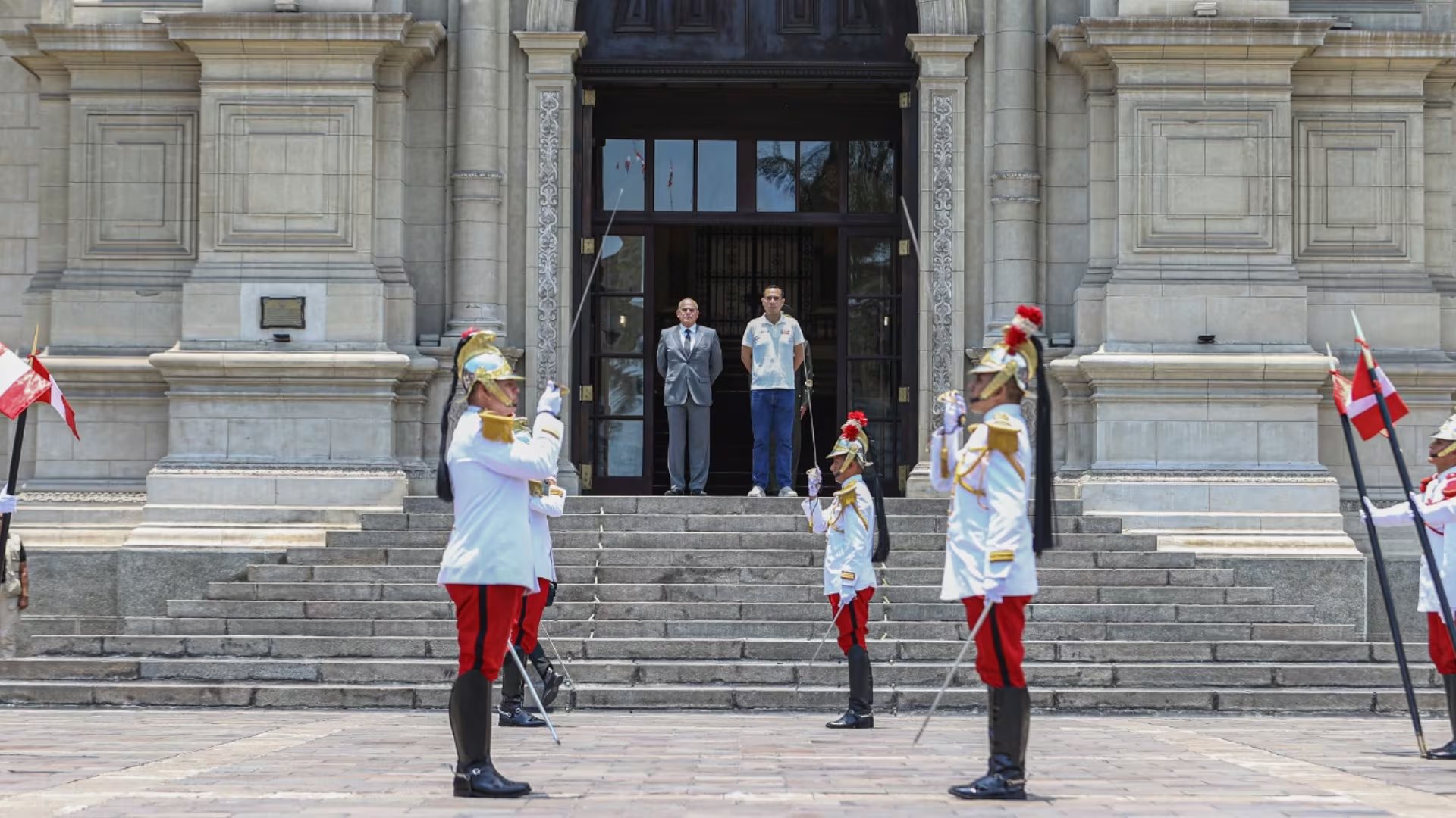 Miembros de la guardia ceremonial, ataviados con uniformes distintivos, realizan una formación frente a un imponente edificio mientras dos hombres los observan desde los escalones principales.