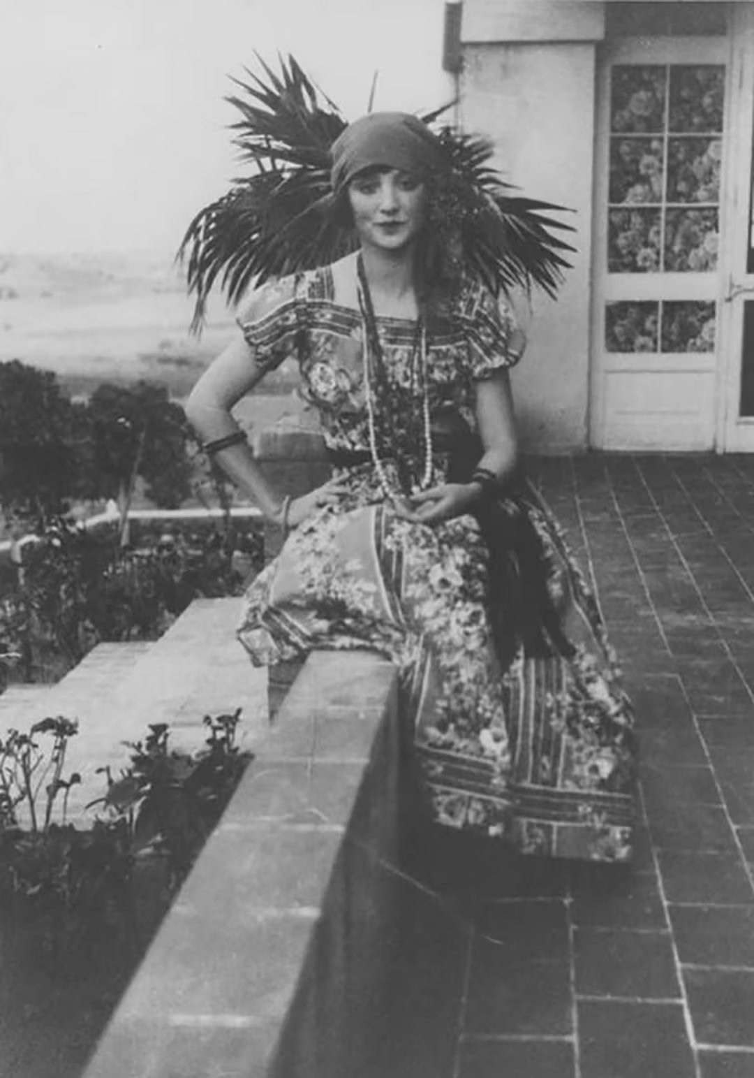Anaïs Nin sentada en un muro, con vestido estampado, tocado y collares largos, imagen de su estilo bohemio y enigmático

