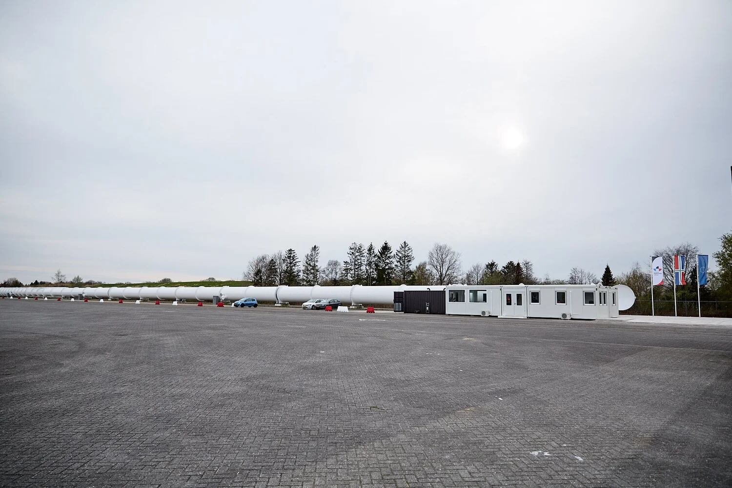 La pista de prueba del Centro Europeo Hyperloop se extiende con su tubo blanco, acompañada de edificios modulares y algunos vehículos en el área de estacionamiento. (Foto: Centro Europeo de Hyperloop)