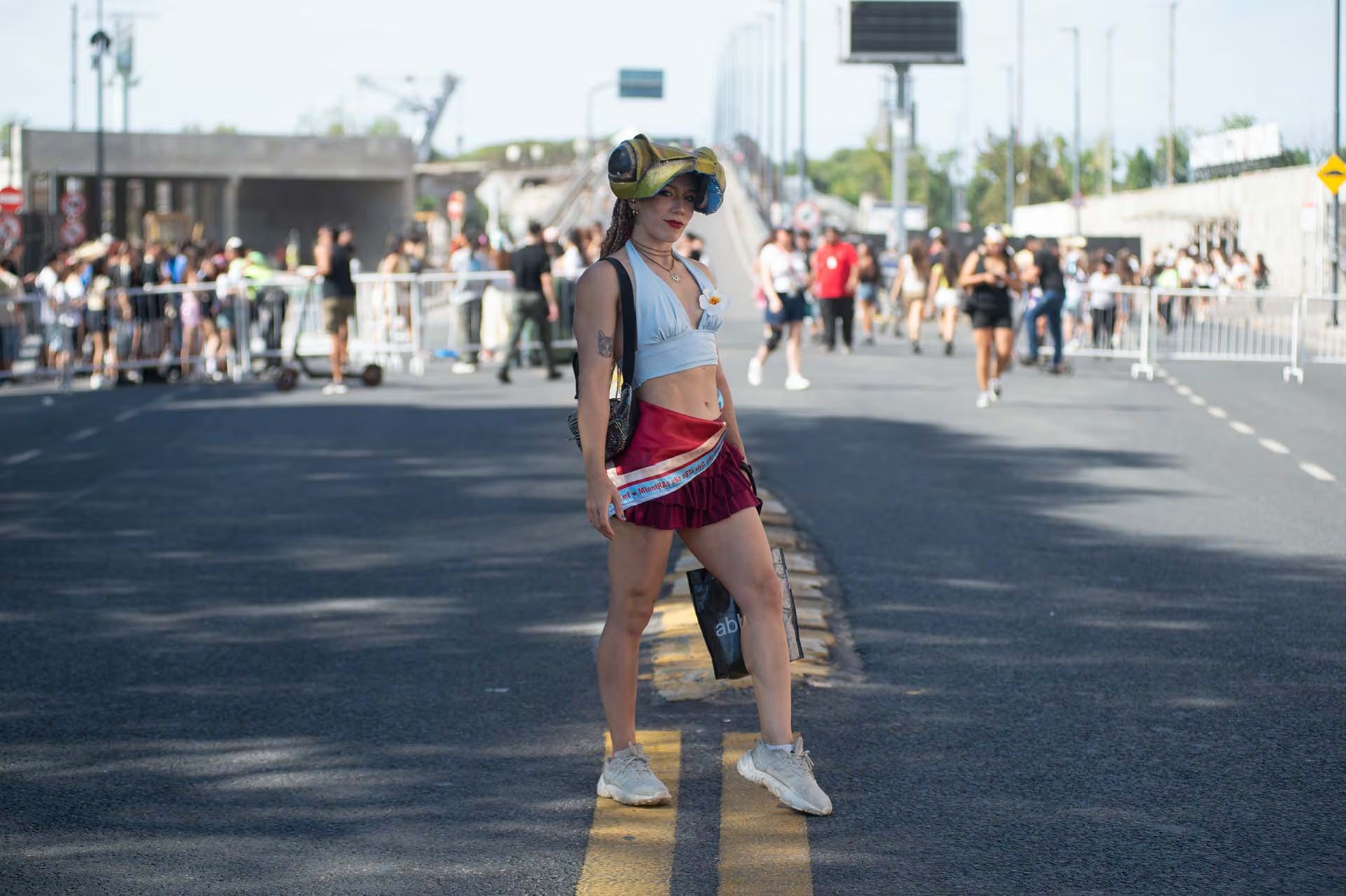 Una joven posa en el centro de una avenida, con la multitud y la estructura del estadio desenfocadas en el fondo. Lleva una falda corta roja, un top celeste con una flor azul y una gorra decorada con grandes ojos de rana, un accesorio que remite a la estética del artista (Jaime Olivos)