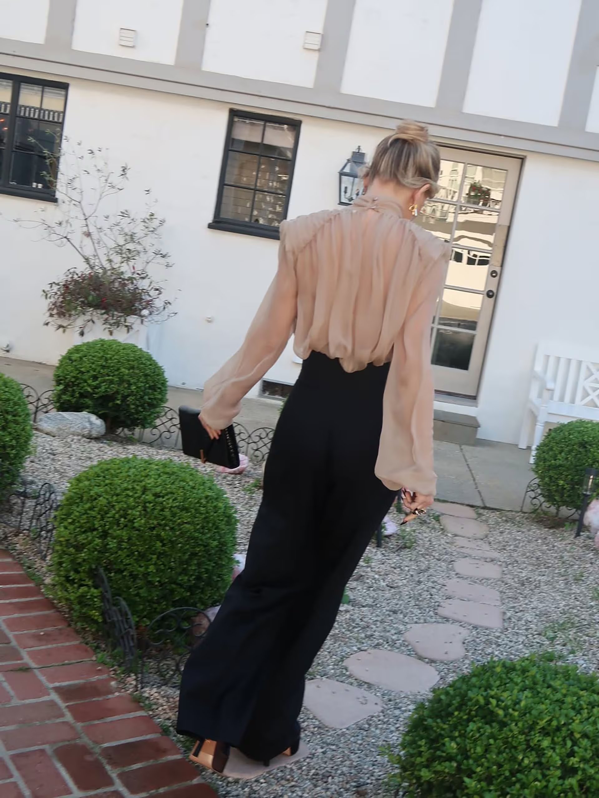 Kate Hudson, vestida elegantemente con una blusa beige y pantalones negros, camina por un sendero de piedra en un jardín, reflejando su continuo éxito.