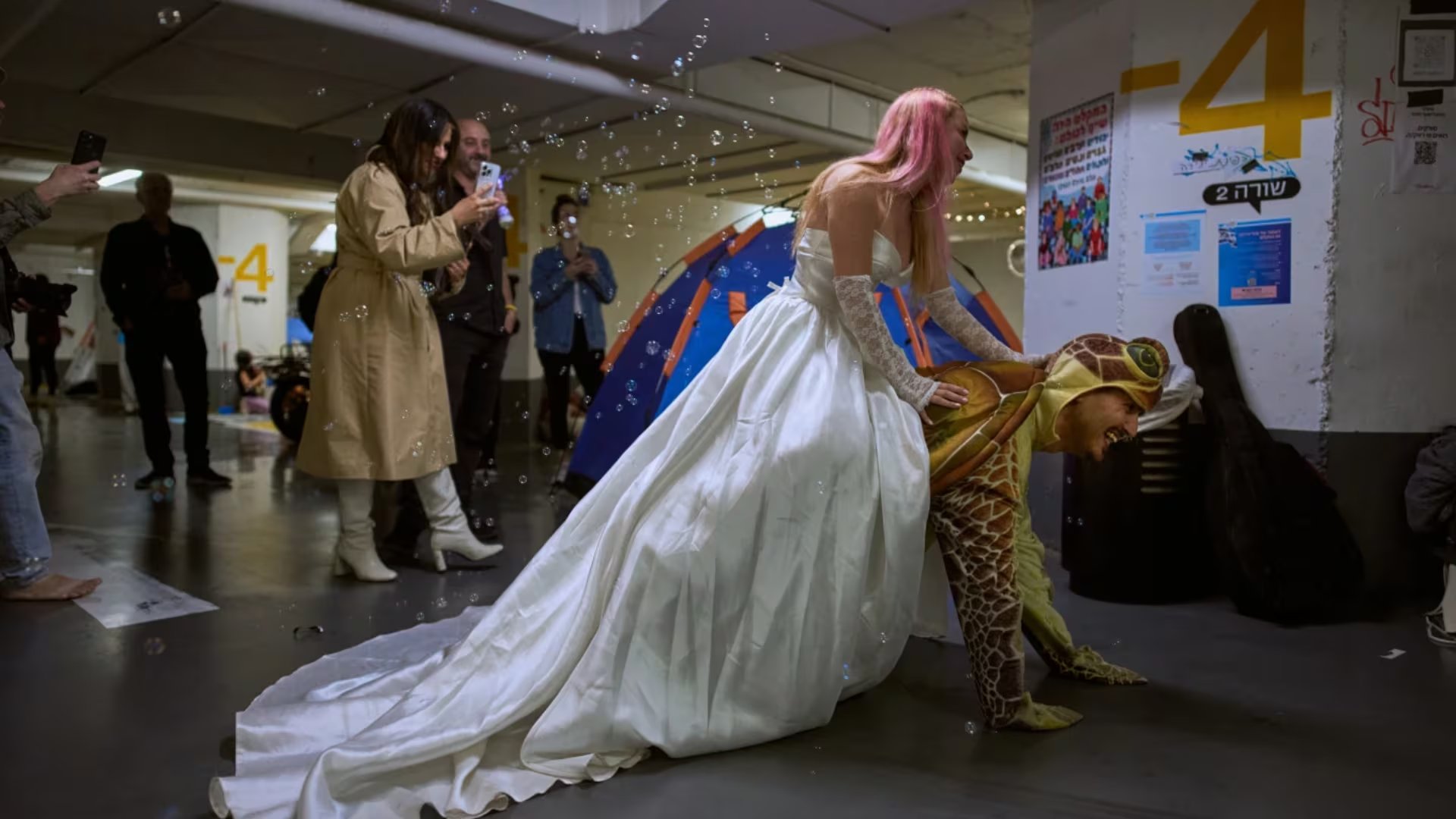 Una mujer vestida de novia viaja a lomos de un hombre disfrazado de tortuga durante un evento para solteros en un estacionamiento subterráneo utilizado como refugio contra posibles ataques con misiles iraníes en Tel Aviv, Israel, el martes 17 de marzo de 2026 (AP/Oded Balilty)