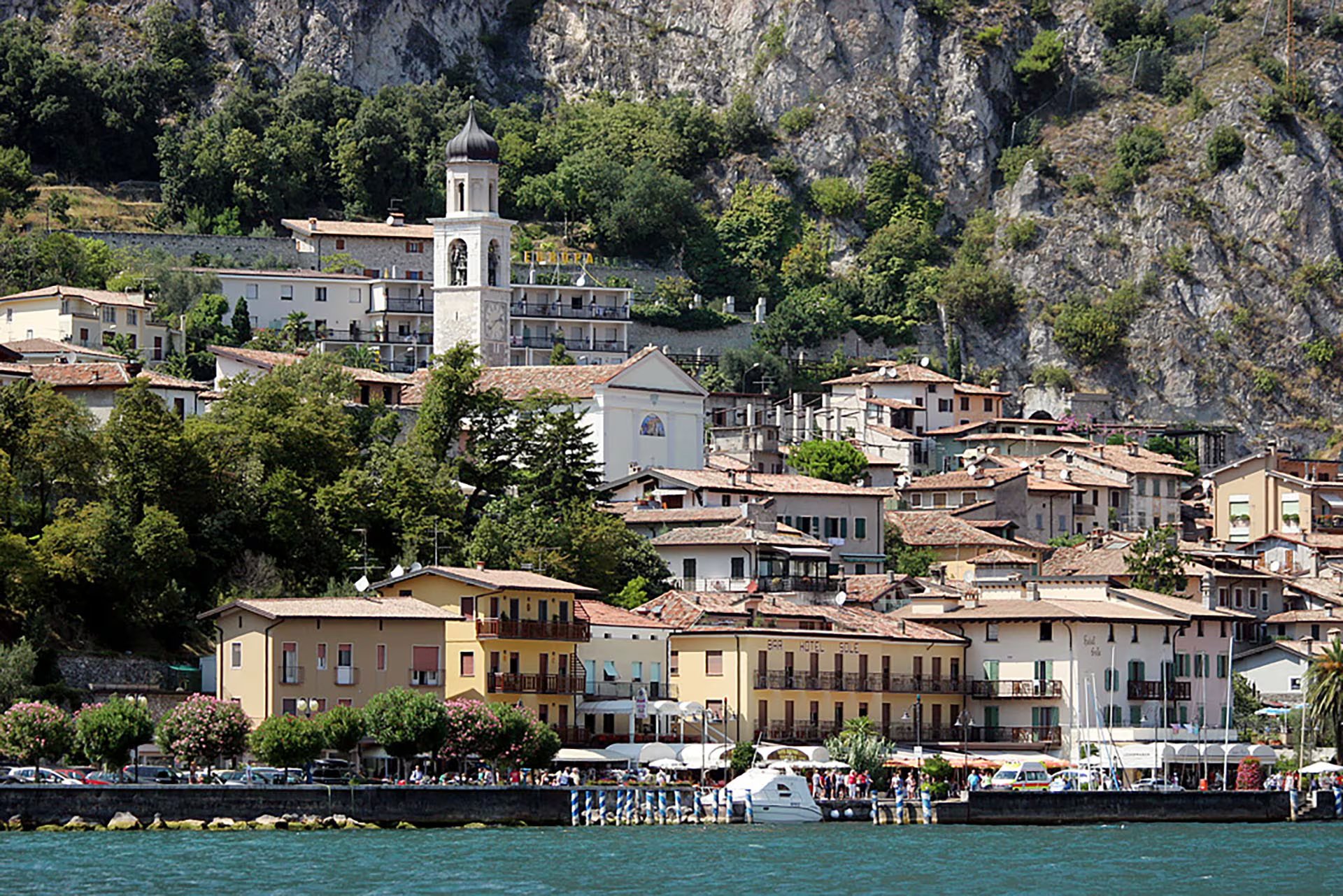 Un pintoresco pueblo italiano se extiende a orillas del Lago de Garda, con casas de tejados rojos y una iglesia destacada, rodeado por montañas frondosas (Wikipedia)