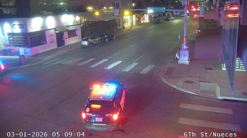 Un vehículo policial con las luces de emergencia encendidas se detiene en una calle urbana iluminada por la noche, indicando una posible intervención o vigilancia en la intersección de 6th St y Nueces. (City of Austin)