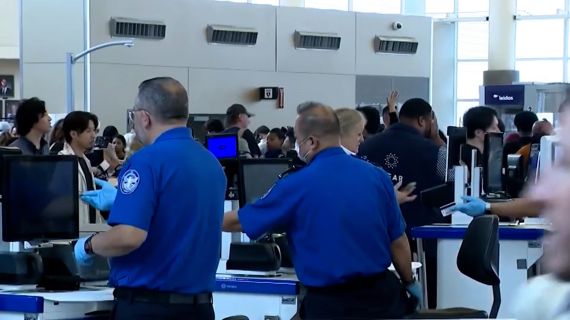 Oficiales de seguridad aeroportuaria con guantes azules asisten a viajeros en un punto de control en un aeropuerto concurrido de Estados Unidos, monitoreando el flujo de pasajeros y equipaje.