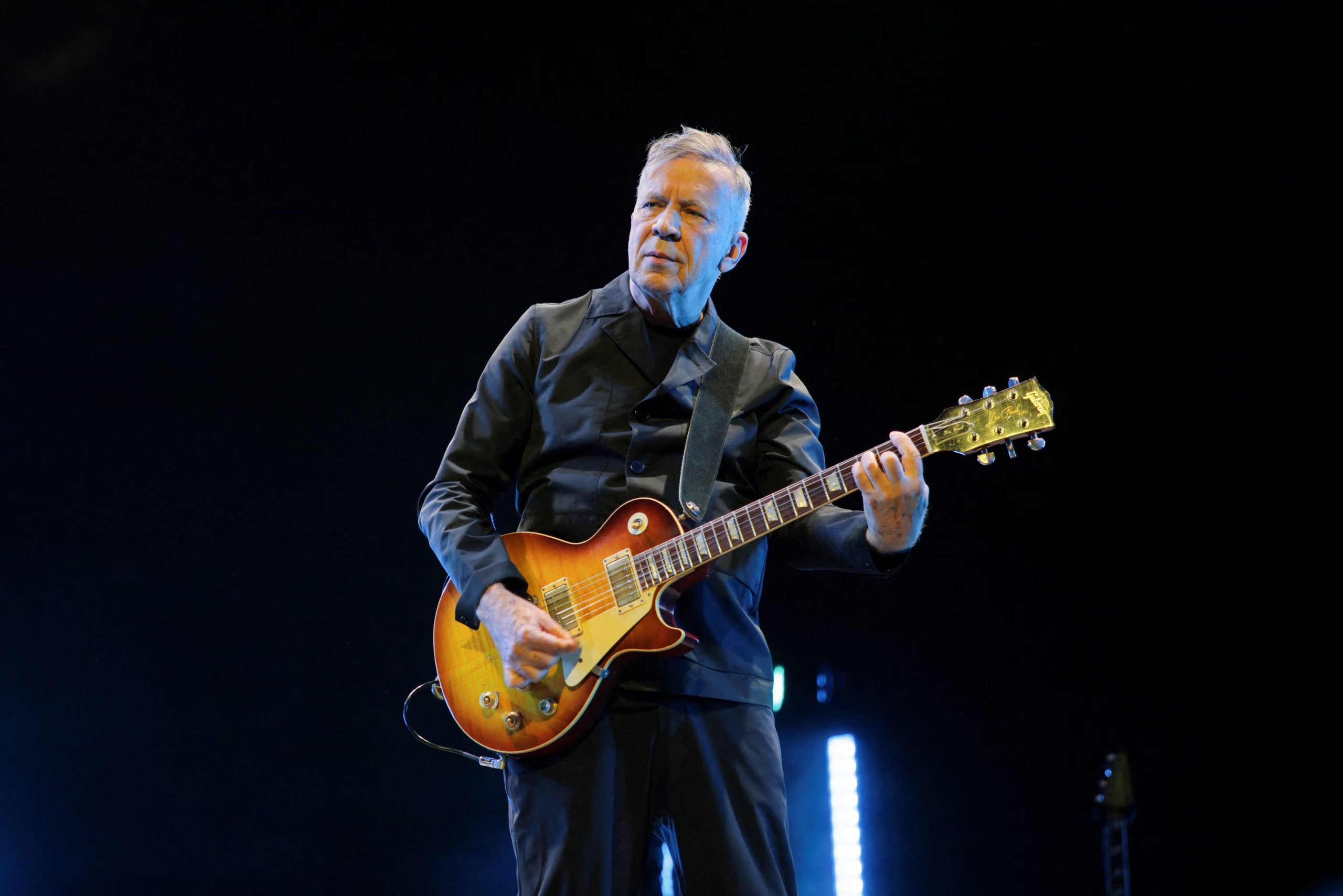Boc Mothersbaugh apostó por la elegancia con un saco cruzado negro y pantalón a juego, complementando con una guitarra eléctrica marrón de correa oscura en escena (REUTERS/Daniel Cole)