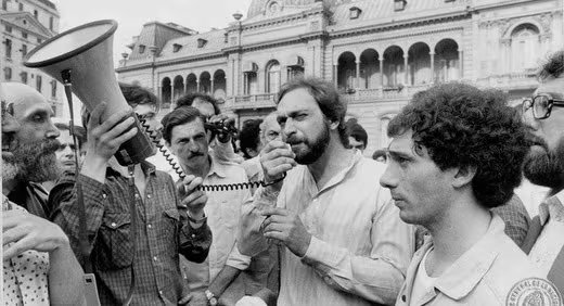 El actor Luis Brandoni habla por un micrófono rodeado de personas, incluyendo a Eduardo Blanco, durante una manifestación pública frente a la Casa Rosada.