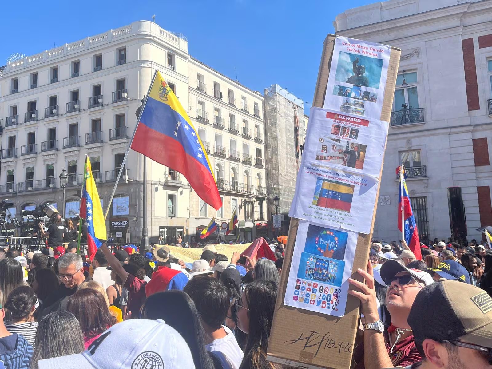 Una multitud de personas se reúne en una plaza, portando banderas de Venezuela y pancartas con mensajes políticos bajo un cielo azul despejado.