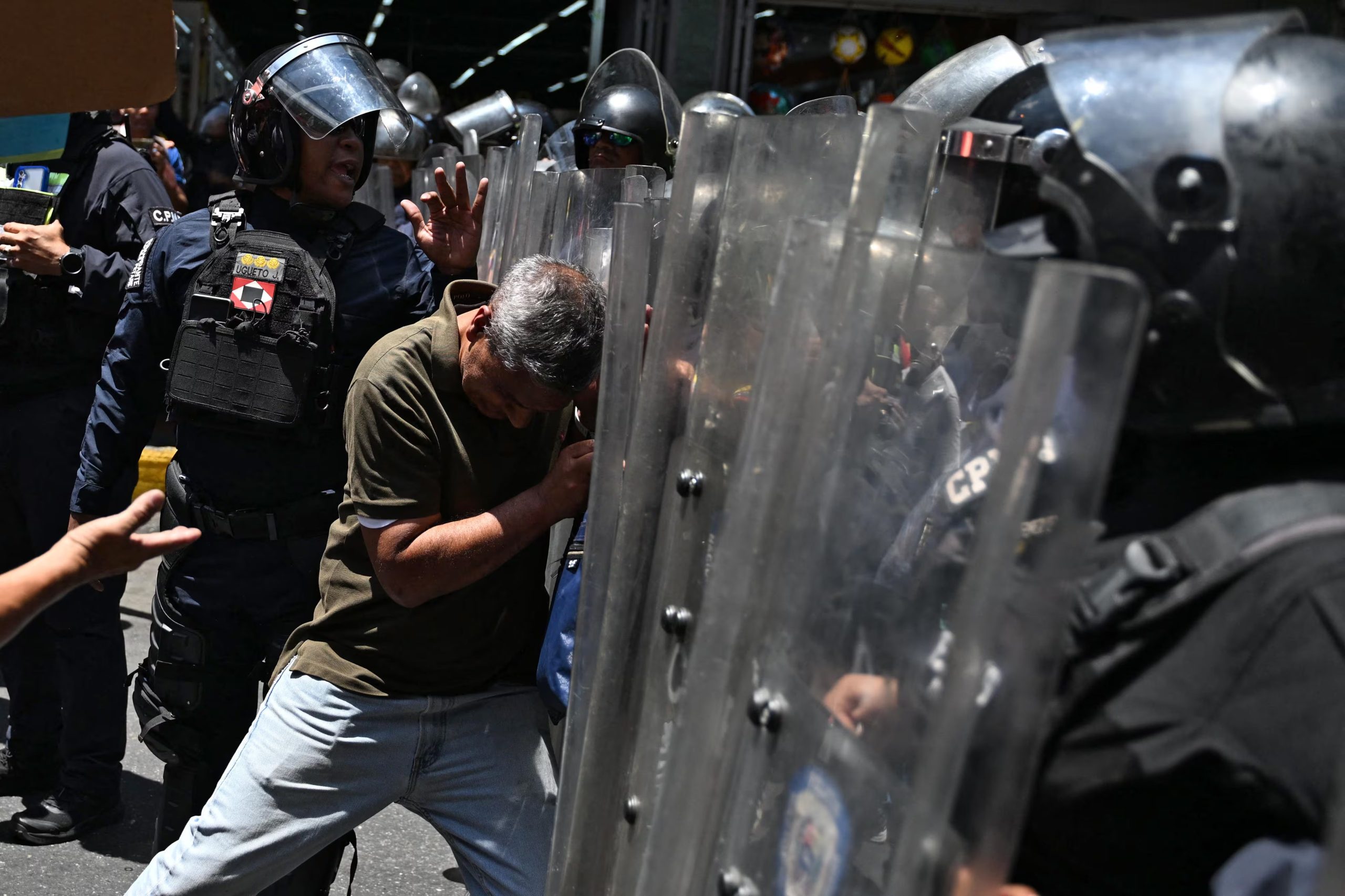 Un hombre empuja con fuerza contra un muro de escudos transparentes sostenidos por policías antidisturbios durante un tenso enfrentamiento en la calle.