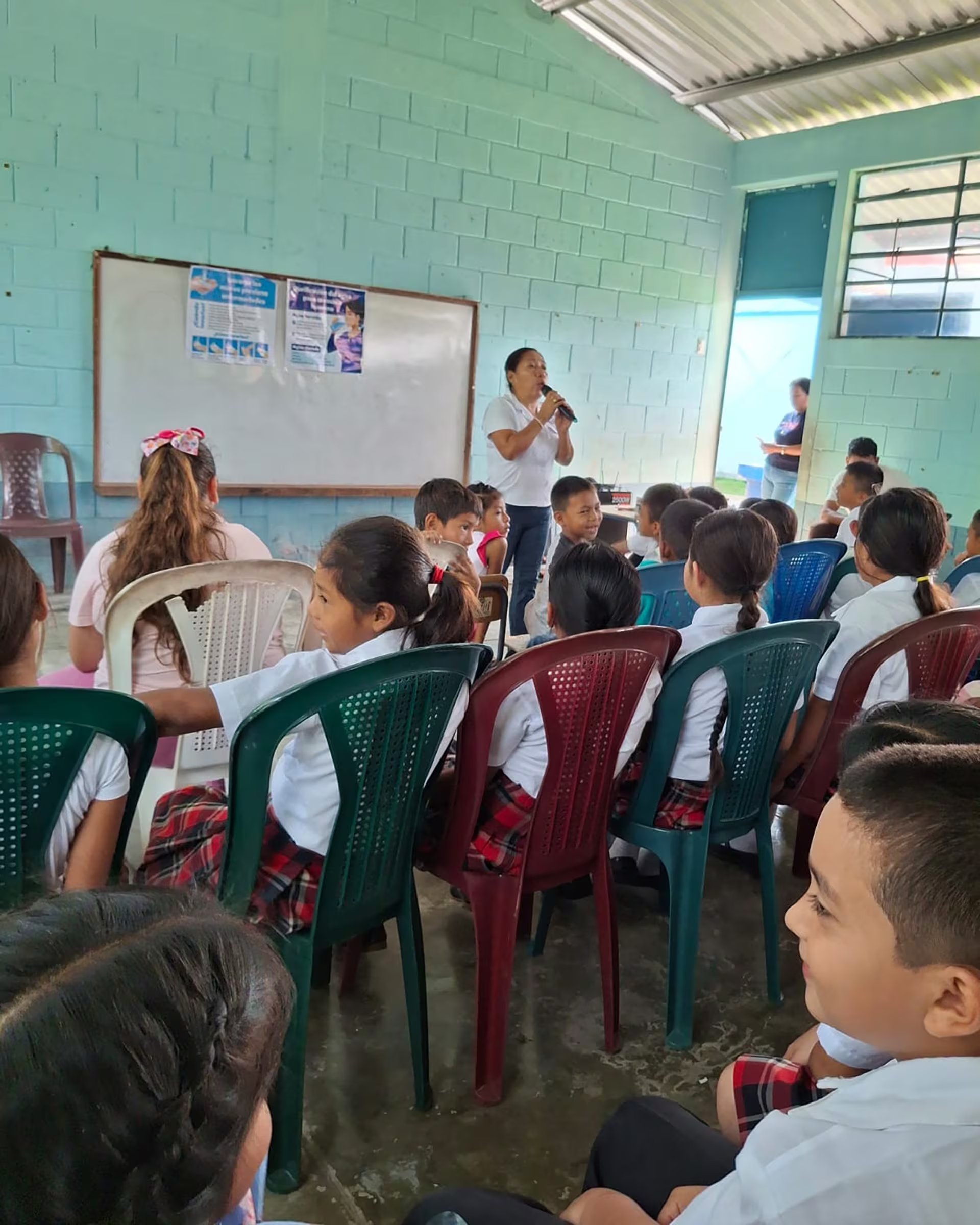 Una maestra imparte una charla educativa a un grupo de estudiantes sentados en un aula, quienes la escuchan atentamente mientras ella usa un micrófono. (Ministerio de Salud y Asistencia Social de Guatemala)