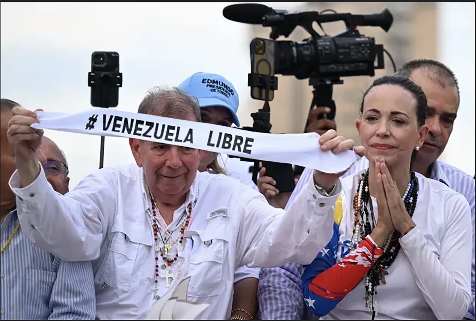 Edmundo González Urrutia y María Corina Machado sostienen una pancarta con el lema '#Venezuela Libre' durante un evento, siendo captados por un equipo de televisión