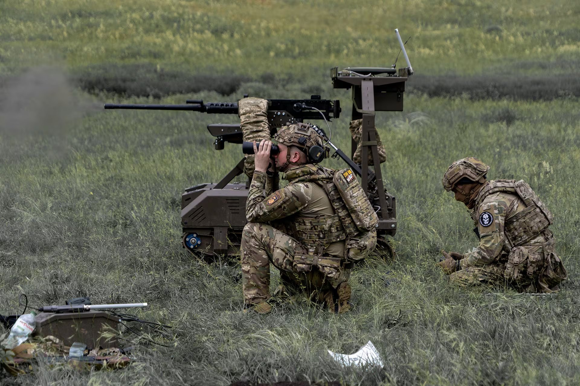 Un soldado ucraniano observa a través de binoculares mientras un sistema de arma robotizada dispara en un campo, reflejando la preparación del país ante la escalada del conflicto con Rusia y la cautela hacia Bielorrusia.