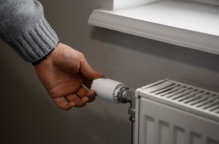 Una mano ajusta el termostato de un radiador blanco para regular la temperatura ambiente, simbolizando el control sobre el sistema de calefacción. (Freepik)