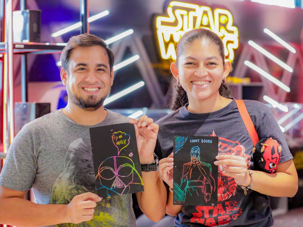 Dos aficionados de Star Wars posan con orgullo sus creaciones de arte para rascar, representando a Darth Vader y el Conde Dooku, frente a un luminoso cartel de la saga. (Foto cortesía)