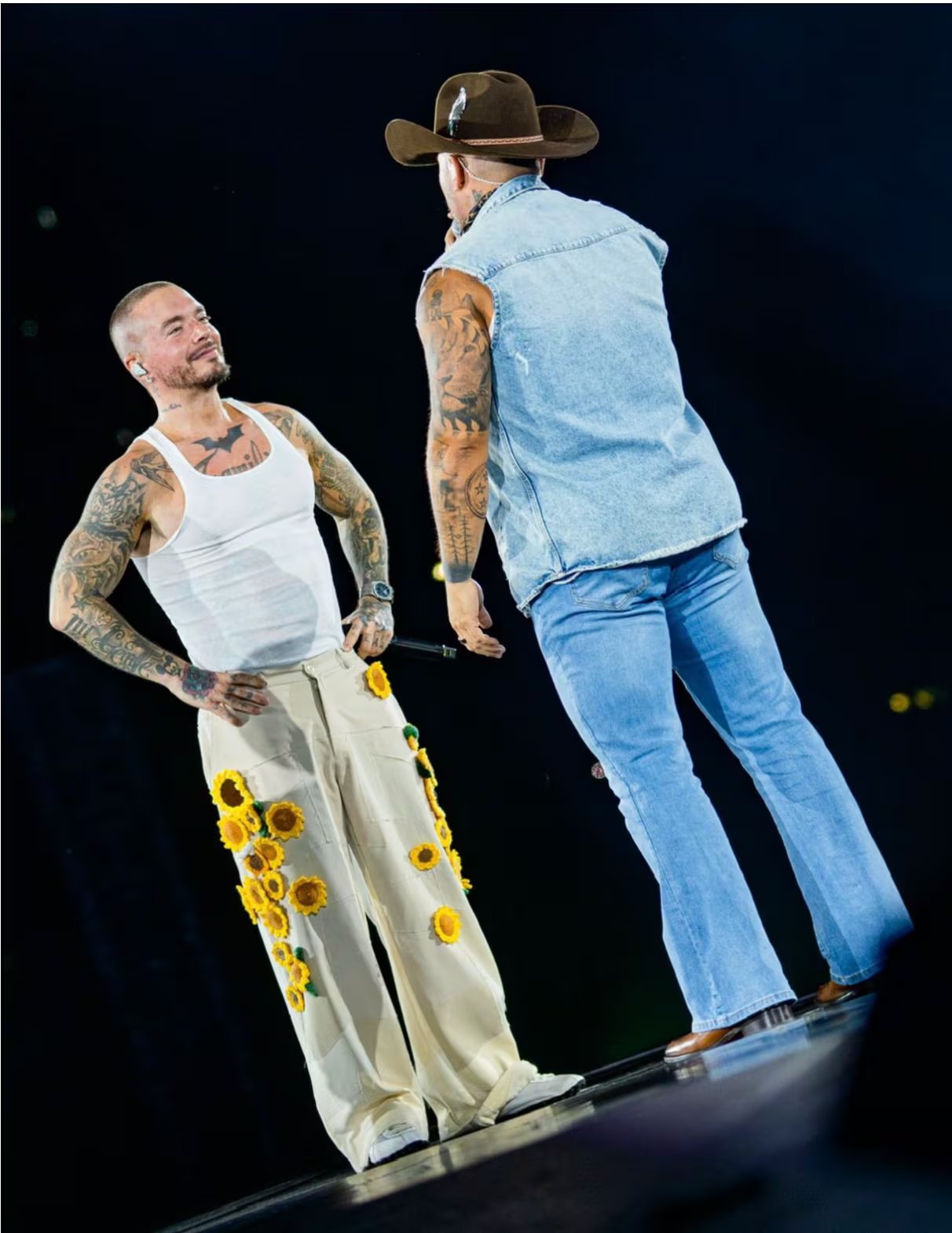 J Balvin con camiseta blanca sin mangas y pantalones crudo decorados con girasoles amarillos, zapatillas blancas y reloj negro, mostrando tatuajes y un estilo desenfadado
