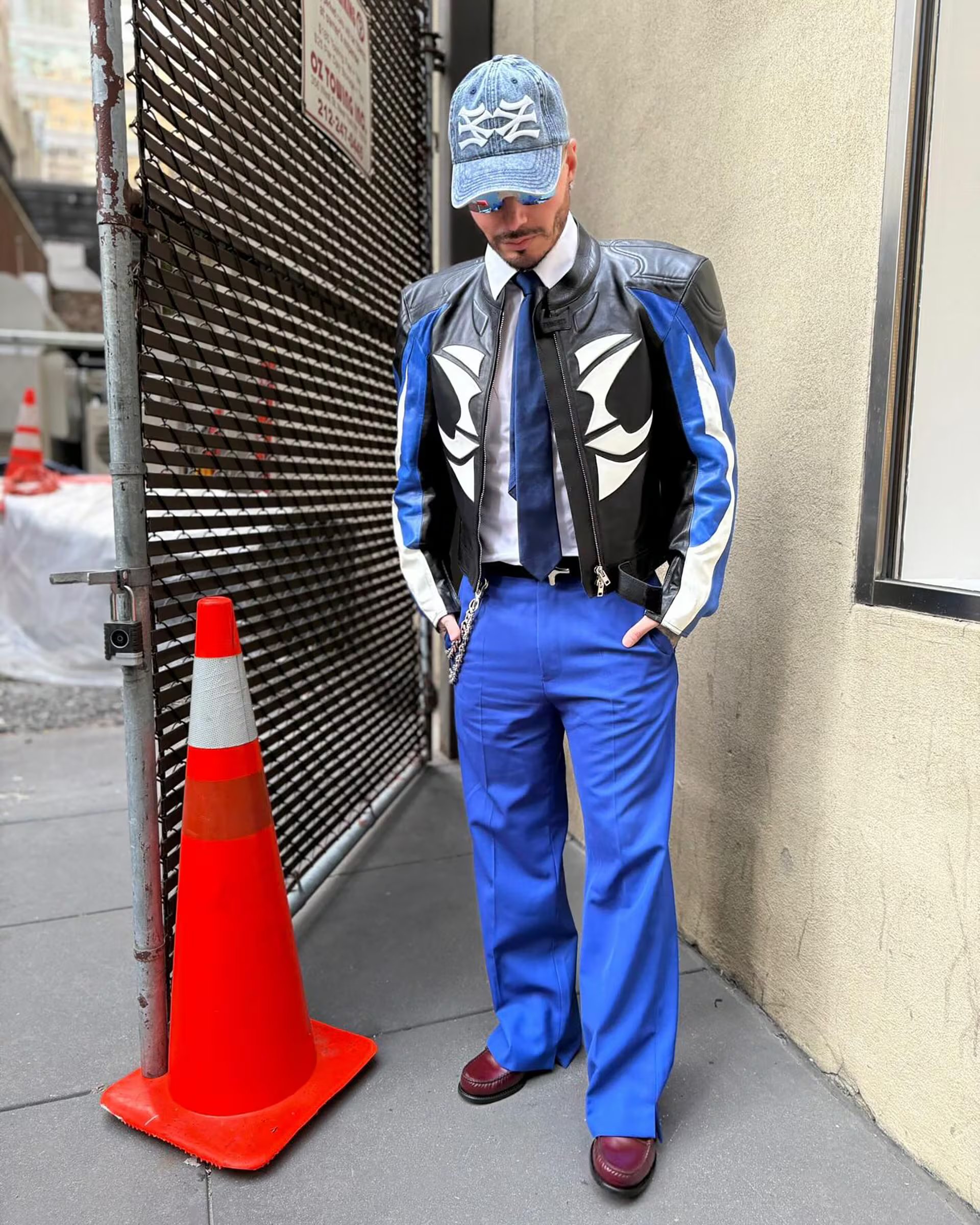 Chaqueta de cuero bicolor negro y azul con detalles blancos, pantalón azul intenso, camisa blanca, corbata negra, gorra vaquera azul y gafas espejadas (@jbalvin)