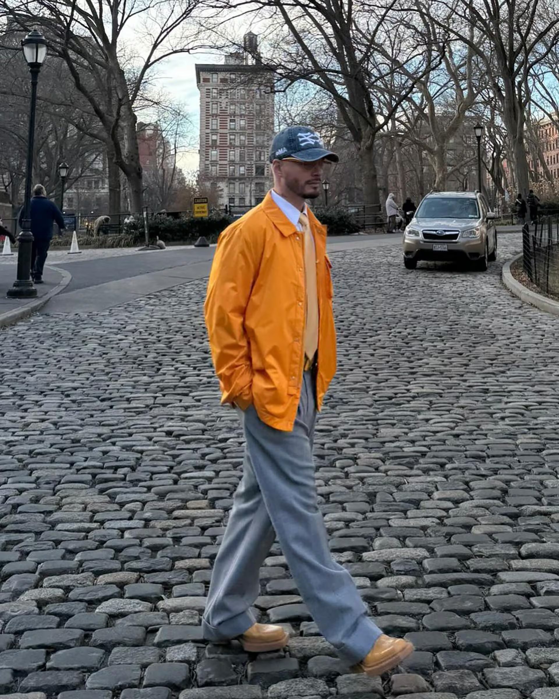 Chaqueta ligera naranja, camisa blanca, corbata amarilla, pantalones grises claros, zapatos beige, gorra vaquera azul y gafas de sol en entorno urbano (@jbalvin)