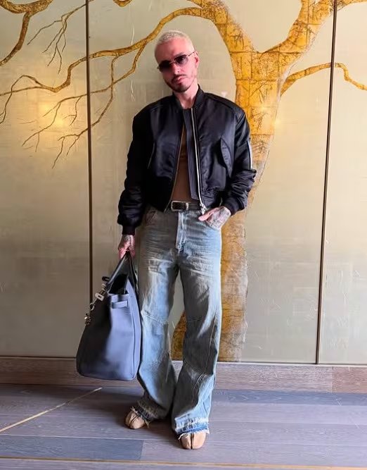 Chaqueta bomber negra, camiseta marrón ajustada, jeans holgados claros, zapatos beige, gafas de sol rosadas y bolso grande gris con cabello rubio claro (@jbalvin/IG)