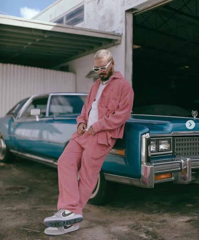 Conjunto de pana rosa con chaqueta y pantalón holgados, camiseta blanca, gafas de sol blancas, cadena plateada y zapatillas deportivas grises apoyado sobre un auto azul (@jbalvin/Instagram)