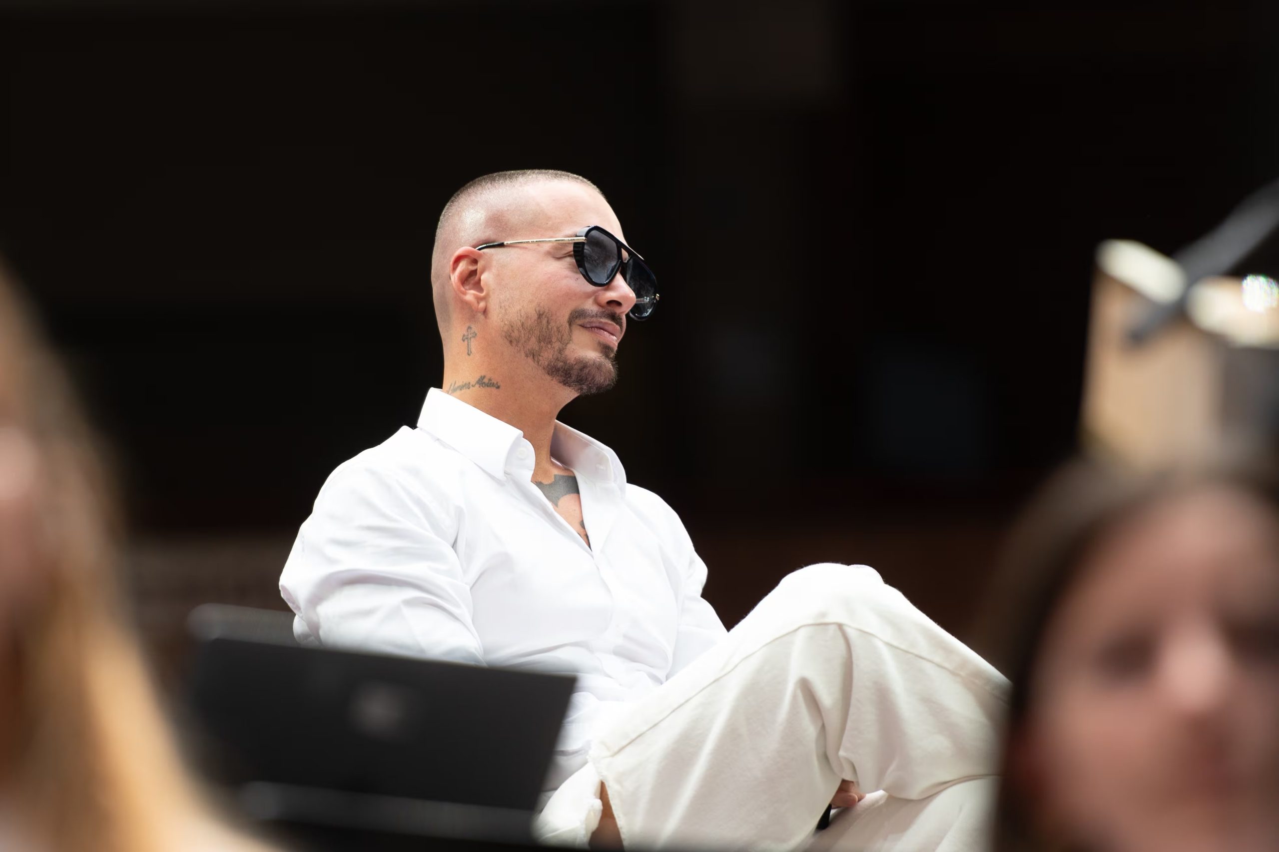 Camisa blanca de algodón y pantalones anchos claros, gafas oscuras grandes y estilismo minimalista en una ocasión formal, sentado de lado y actitud relajada (Vibra en Alta)