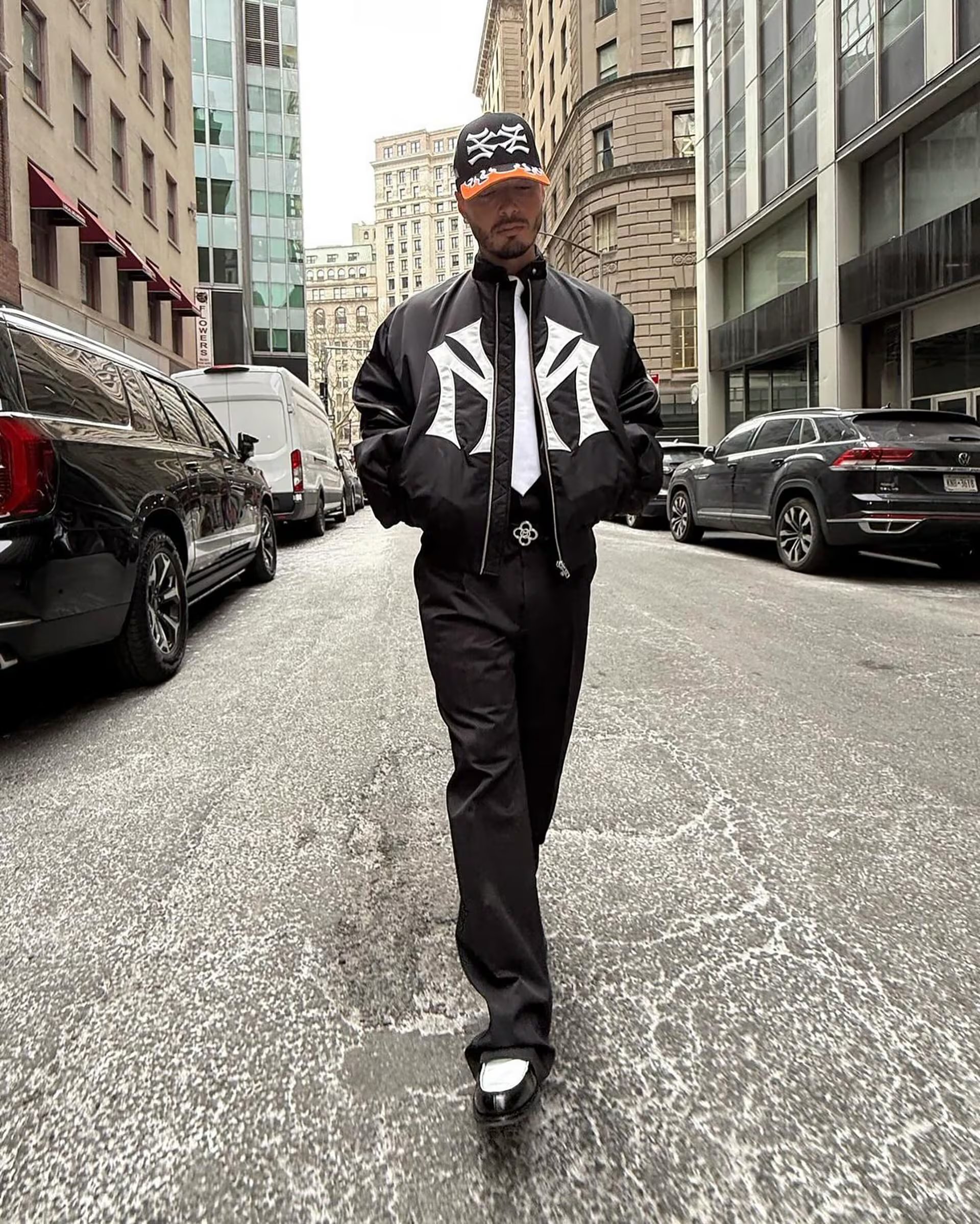 Chaqueta negra con logos blancos, pantalón negro, camisa blanca, corbata negra, zapatos bicolor y gorra negra y naranja en un look urbano y gráfico (@jbalvin)