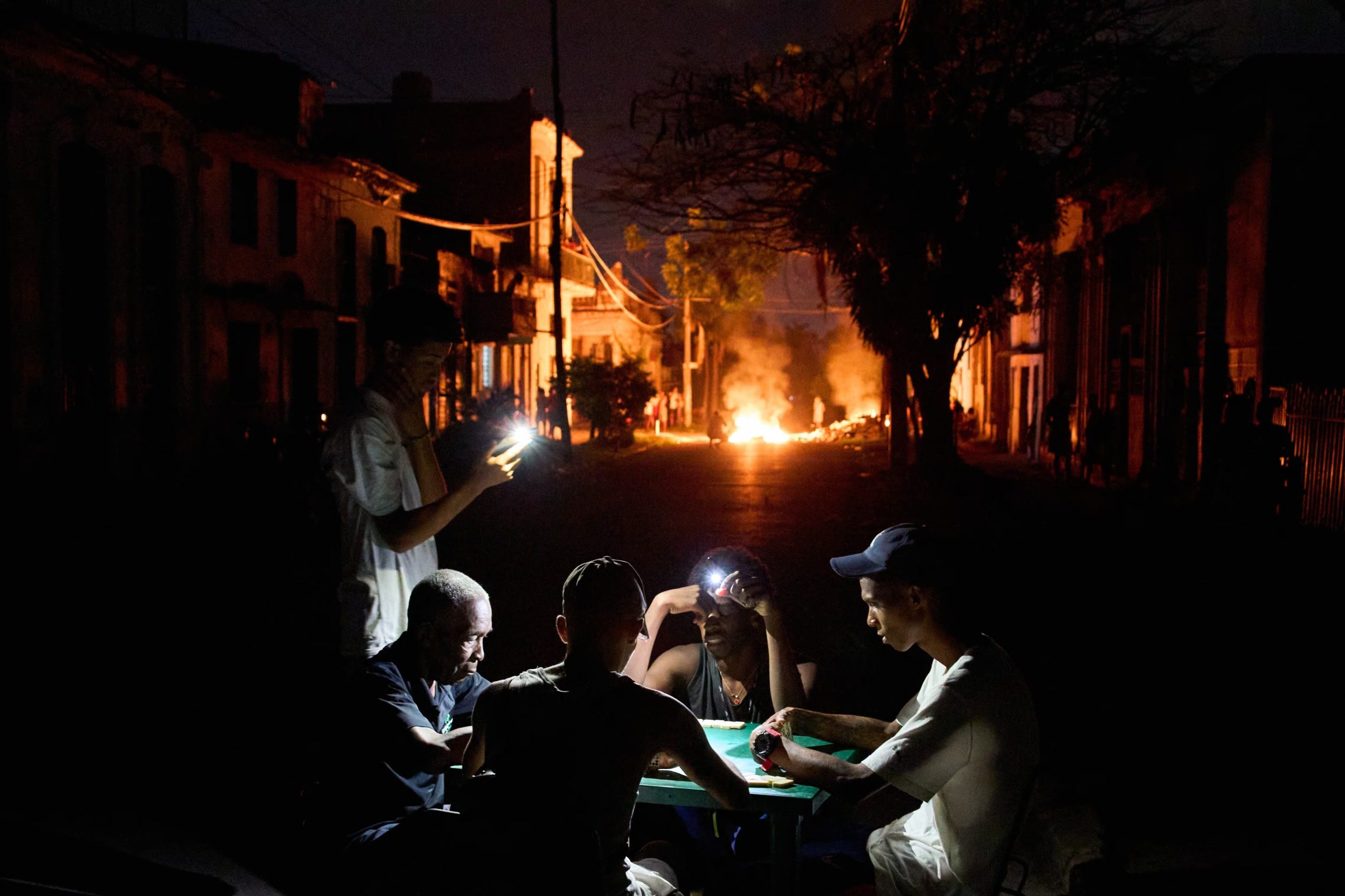 Varias personas se iluminan con sus teléfonos mientras juegan al dominó, al tiempo que arde en una calle de La Habana, Cuba, una hoguera provocada por residentes que protestaban contra los prolongados cortes de luz (Foto AP/Ramón Espinosa)