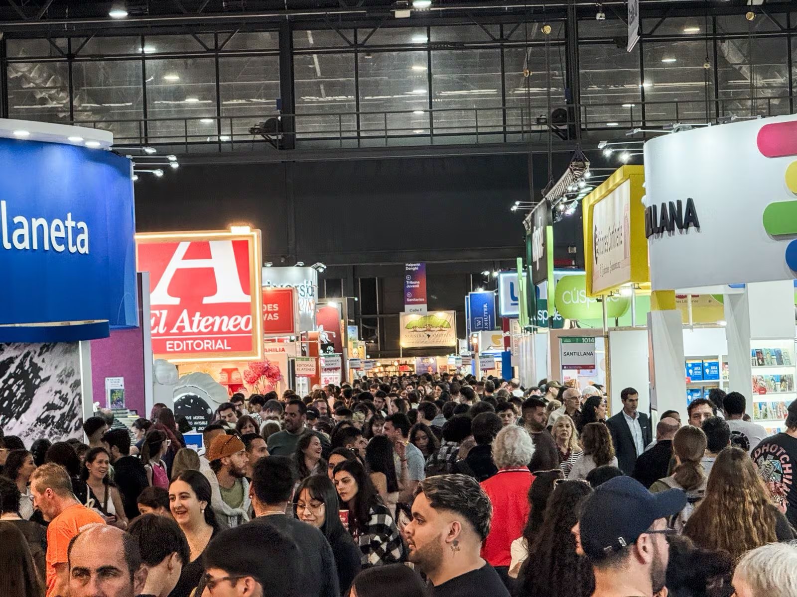 Miles de entusiastas lectores llenan los pasillos de una convención literaria, explorando los diversos stands de editoriales bajo coloridos banners publicitarios.
