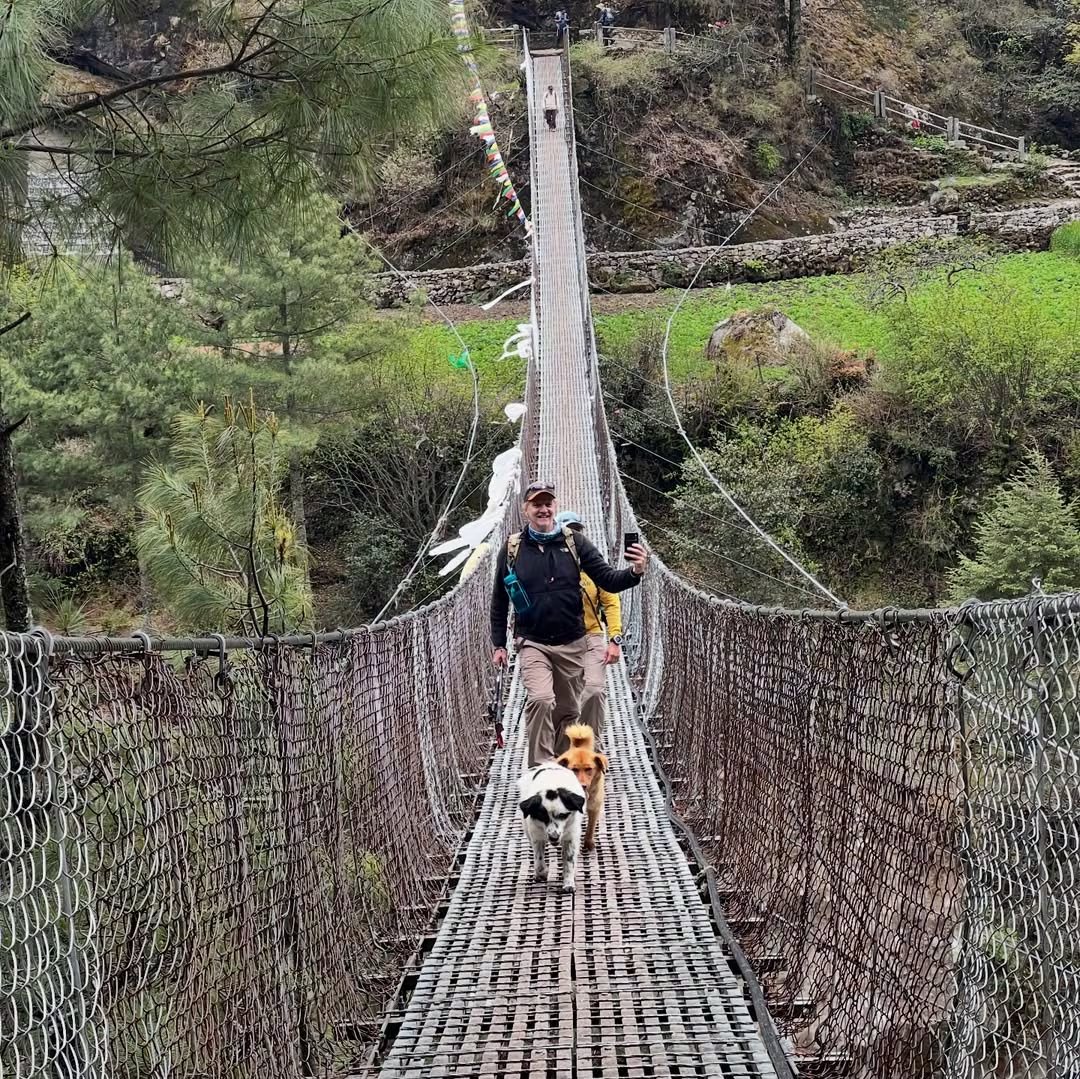 El actor Facundo Arana documenta su expedición al Everest con una selfie junto a dos perros mientras cruza un puente colgante en un paisaje montañoso.
