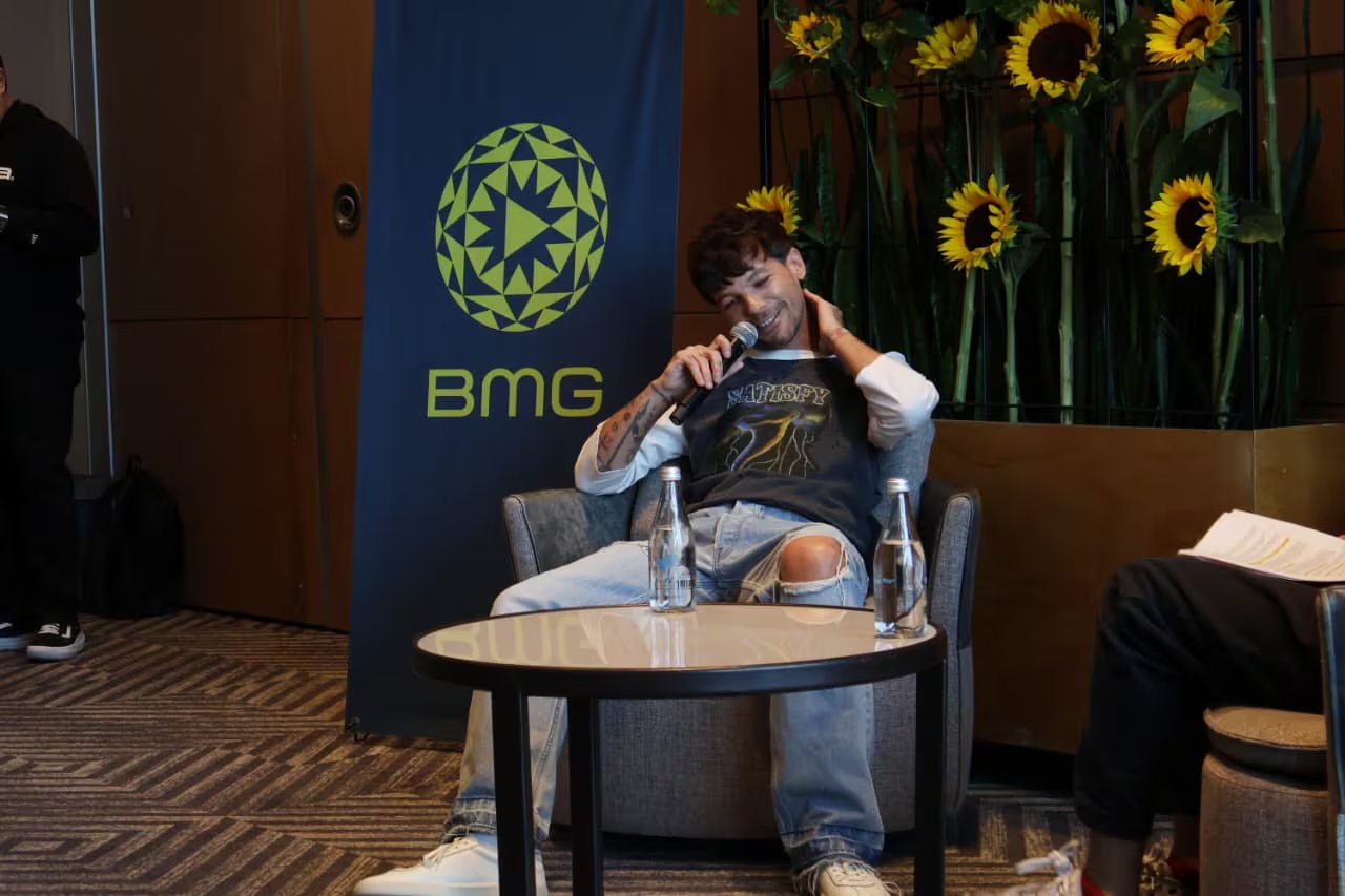 El cantante Louis Tomlinson sonríe y sostiene un micrófono mientras participa en una entrevista, con un banner de BMG y girasoles de fondo. (Selene Miranda)