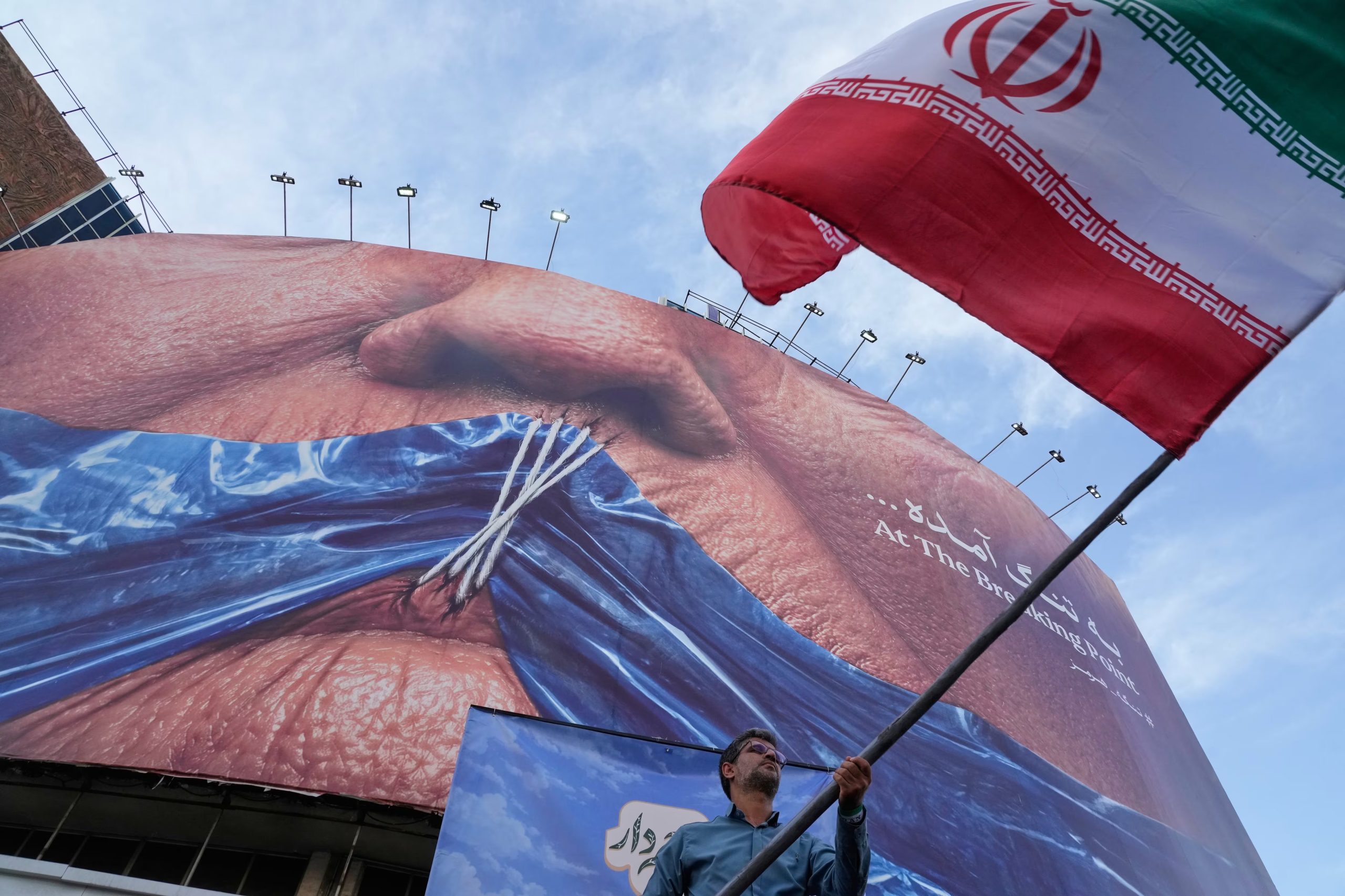 Un hombre ondea una bandera iraní frente a un cartel que muestra el Estrecho de Ormuz y los labios cosidos del presidente Donald Trump, en Teherán el 6 de mayo de 2026. (Vahid Salemi/AP)