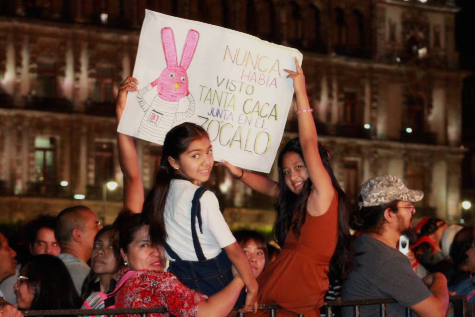 Dos jóvenes fans sonríen mientras sostienen una pancarta creativa con un conejo rosa y un mensaje peculiar en el concierto gratuito de 31 minutos en el Zócalo de la Ciudad de México. (Merary Núñez/ Infobae)