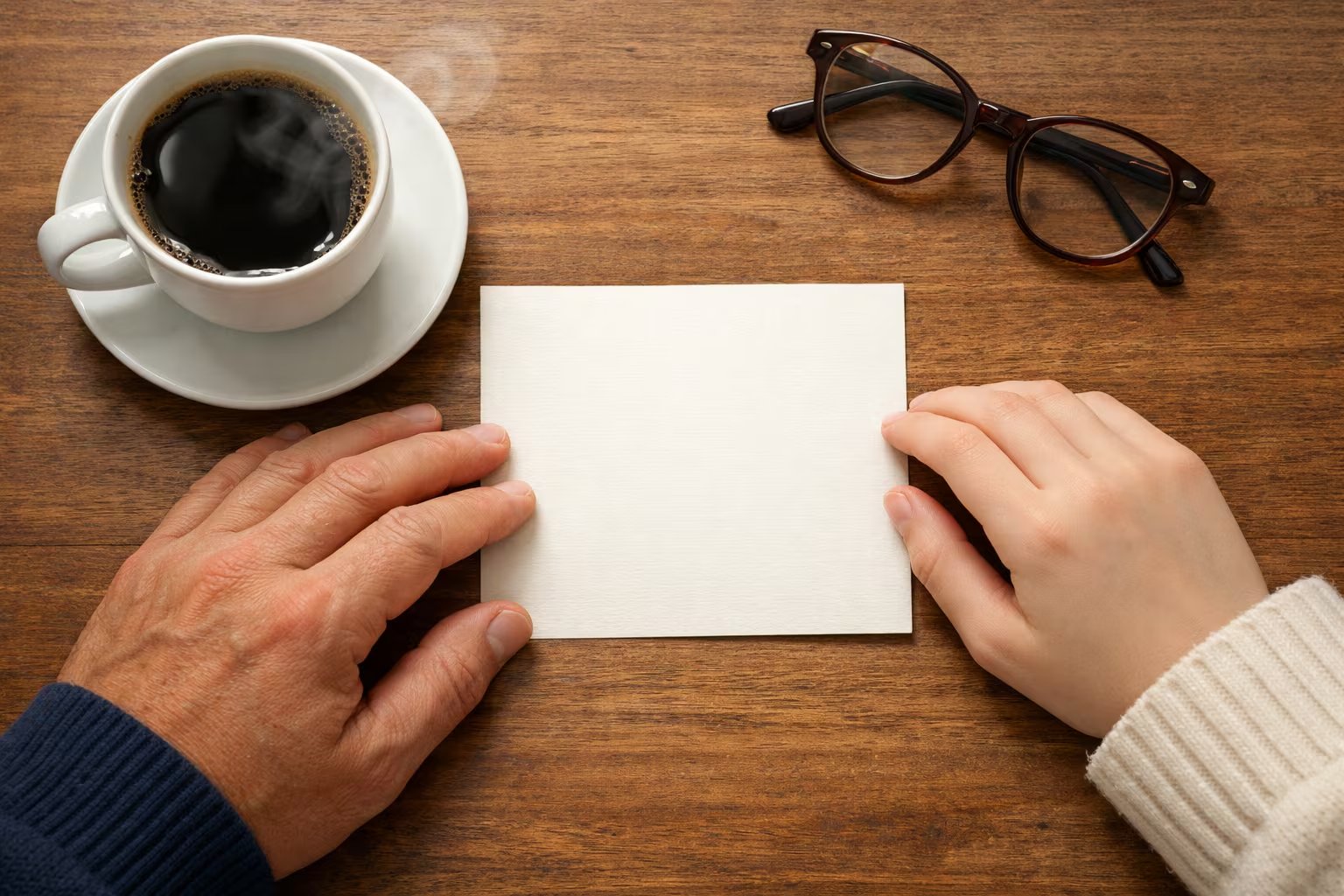 Dos manos, una adulta y una adolescente, sostienen una tarjeta en blanco sobre una mesa de madera, junto a una taza de café humeante y unas gafas, creando una atmósfera de intimidad matutina. (Imagen Ilustrativa Infobae)