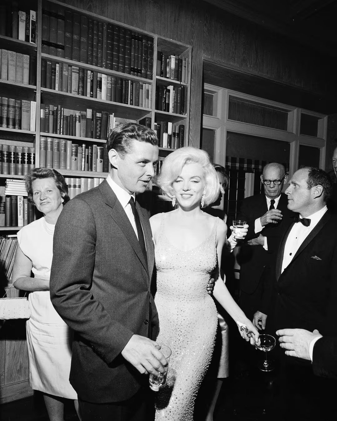 Marilyn Monroe, luciendo su icónico vestido brillante, acompañada por Robert F. Kennedy. Su elegancia combina sensualidad y sofisticación en escotes, tejidos fluidos y conjuntos coordinados (Grosby)