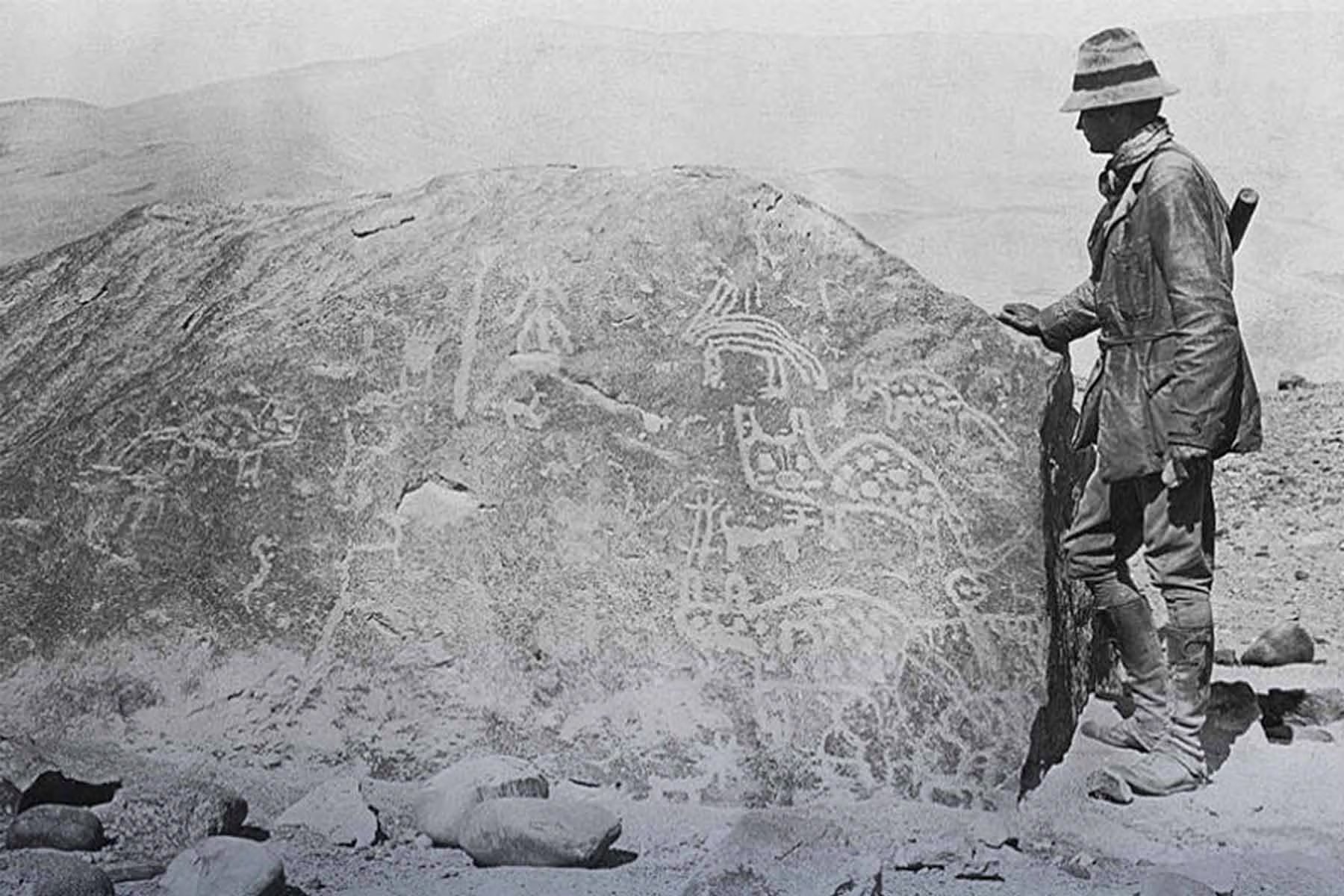 Hiram Bingham III, conocido por sus expediciones en Sudamérica, observa una gran roca cubierta de petroglifos que representan figuras zoomorfas y antropomorfas, testimonio del arte rupestre ancestral.