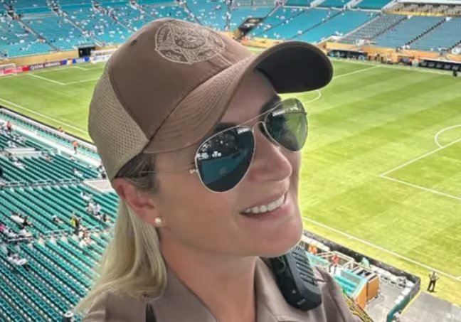 Una mujer con uniforme y gorra, equipada con gafas de sol y radio, sonríe desde una posición elevada con vistas al campo de juego y las gradas de un estadio de fútbol (Captura de video)