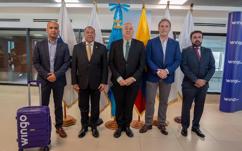 Cinco hombres con atuendo formal se reúnen en un espacio interior, con banderas nacionales de fondo y un cartel de la aerolínea Wingo junto a una maleta morada. (Ministerio de Comunicaciones, Infraestructura y Vivienda)