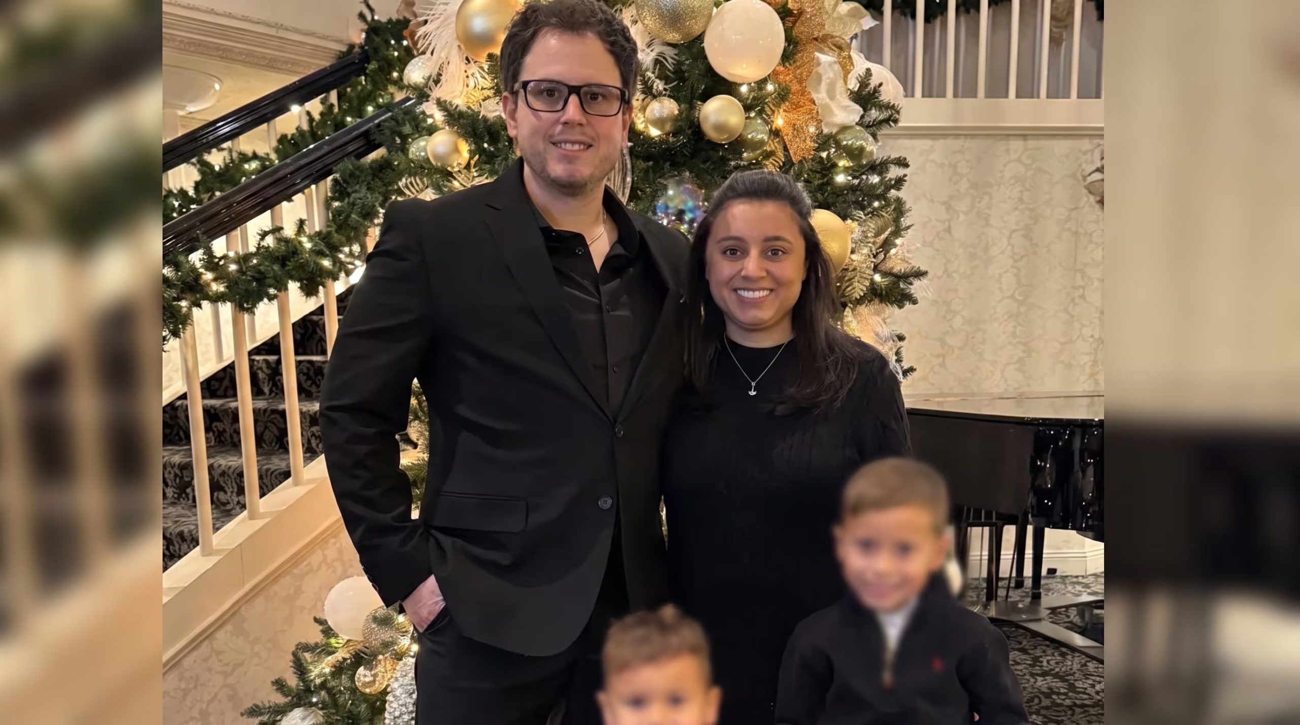 Una pareja posa sonriente frente a un árbol de Navidad con ornamentos dorados y una escalera decorada, con niños difuminados y un piano de cola al fondo. (Cortesía de Nick Nabozny)