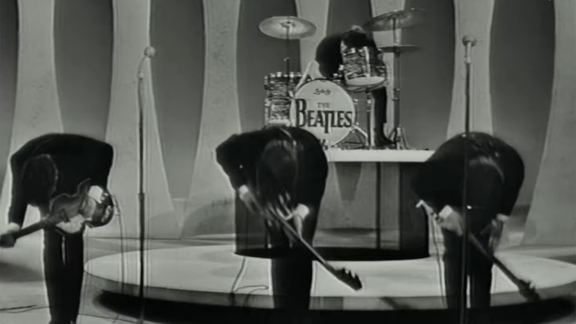 Los miembros de The Beatles se inclinan ante la audiencia después de su actuación, con la batería de la banda visible al fondo en este momento histórico en blanco y negro (Captura de video)