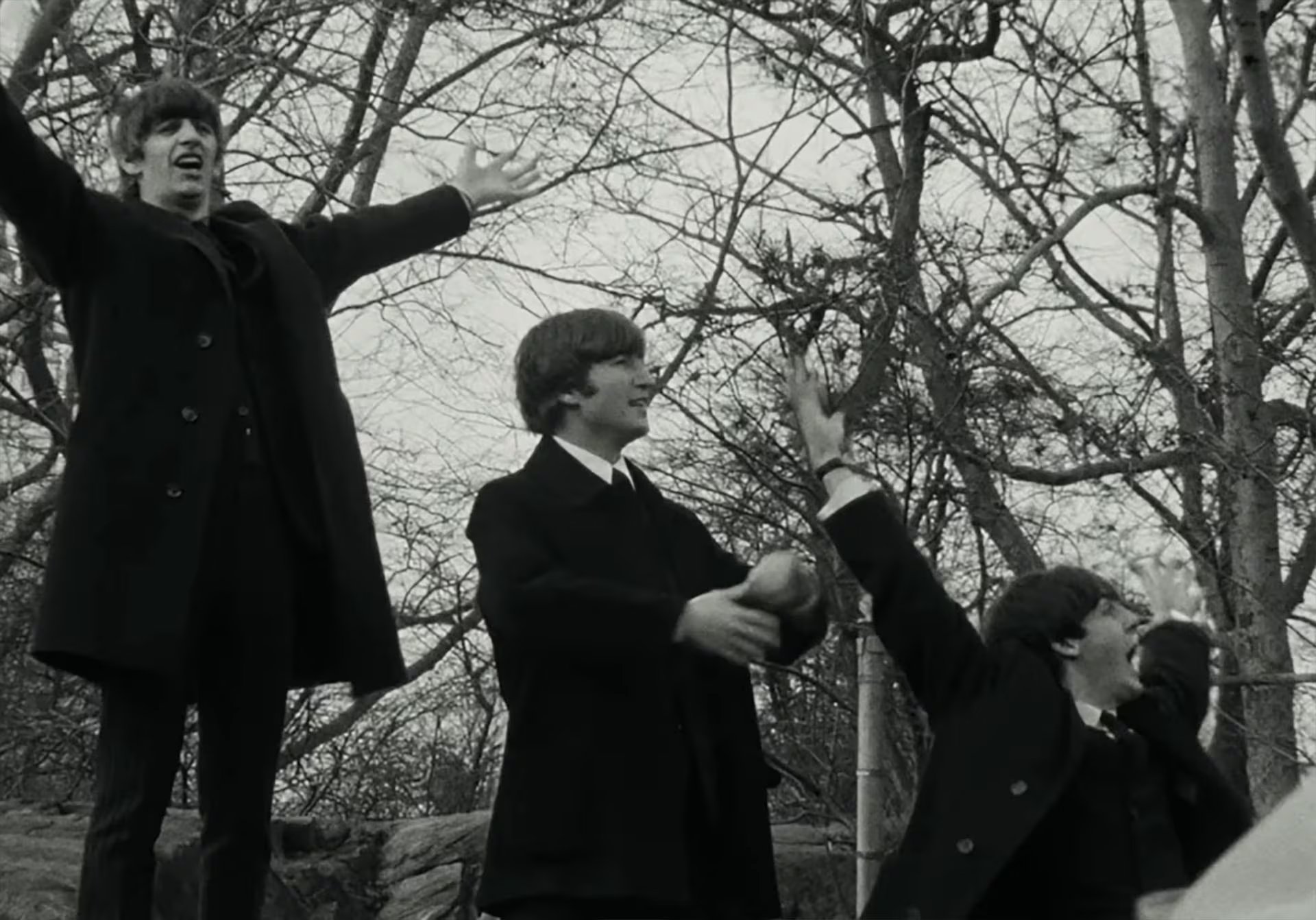 Los miembros de The Beatles, Ringo Starr, John Lennon y George Harrison, posan y gesticulan con los brazos levantados al aire en un parque arbolado durante su primera visita a Nueva York (Captura de video)