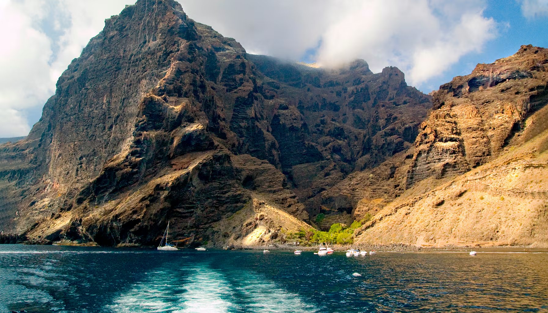 Los acantilados del Barranco de Masca en Tenerife flanquean las aguas azules del océano, donde se observan varias embarcaciones cerca de la costa.