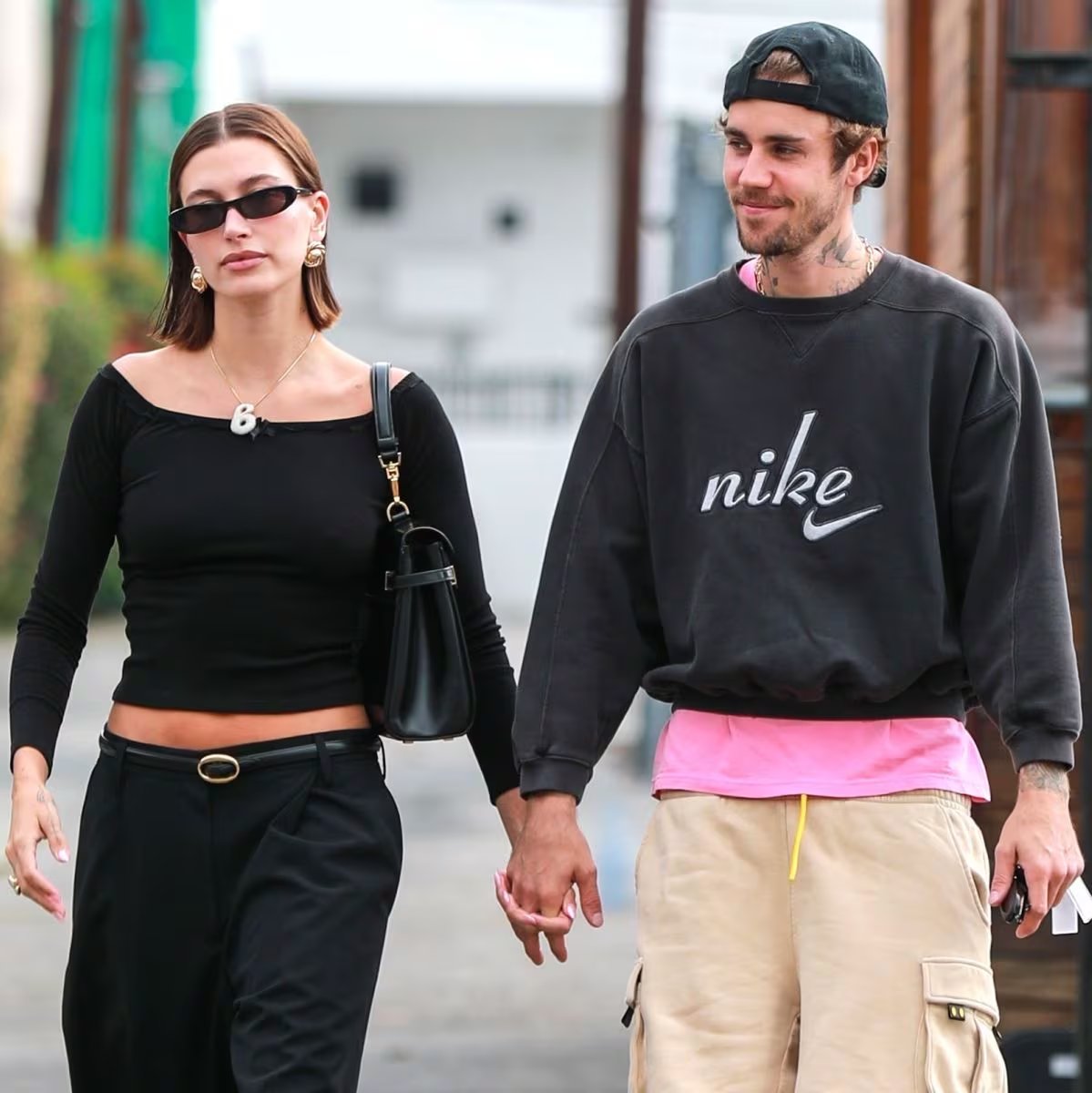 Hailey Bieber arma un conjunto negro de sastrería con accesorios dorados, y Justin Bieber lo cruza con sudadera negra, cargo beige, gorra negra y camiseta rosa (Backgrid/The Grosby Group)