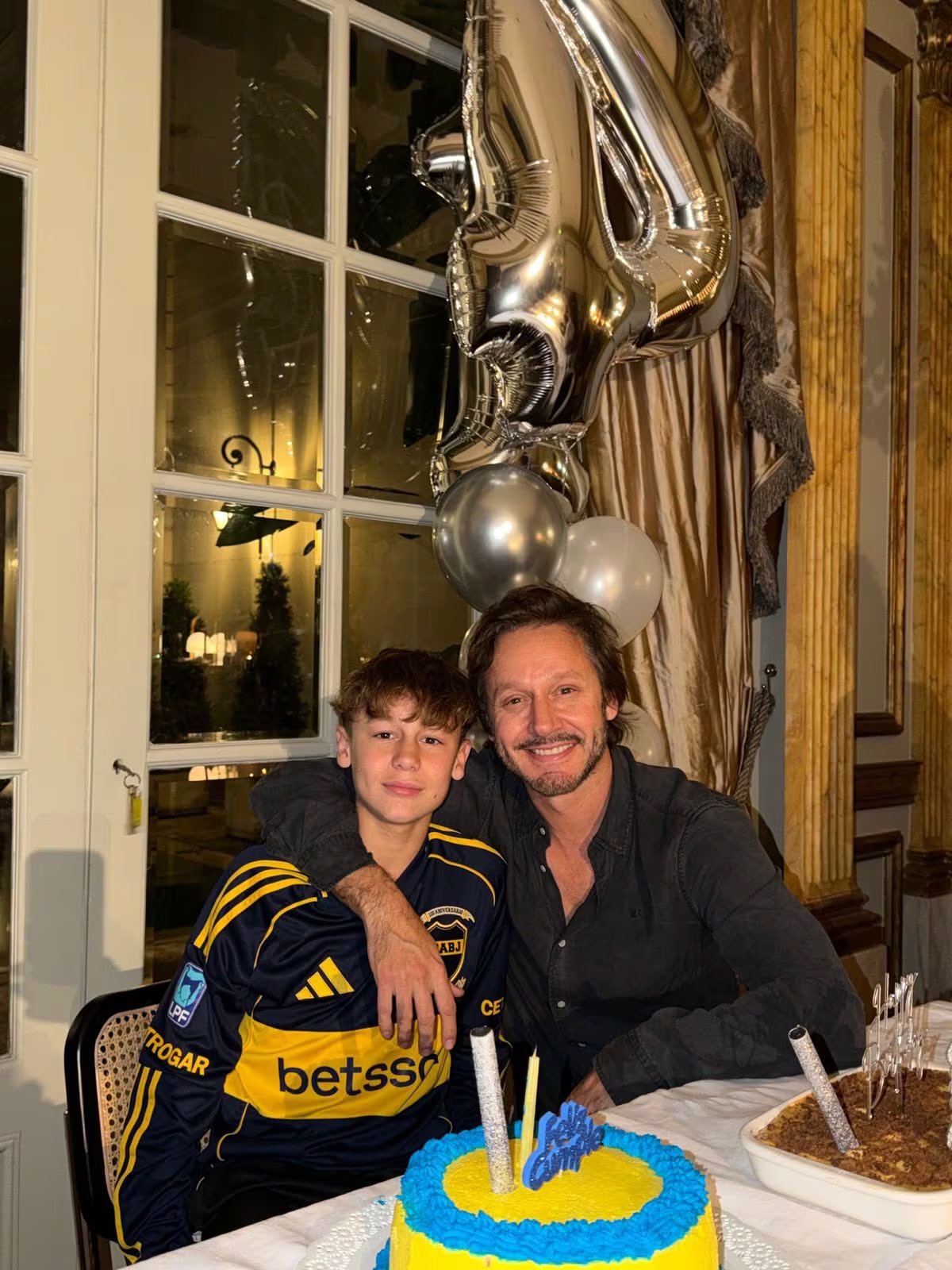 Benjamín Vicuña posa sonriente junto a su hijo, que luce una camiseta de fútbol, frente a una torta de cumpleaños y globos festivos