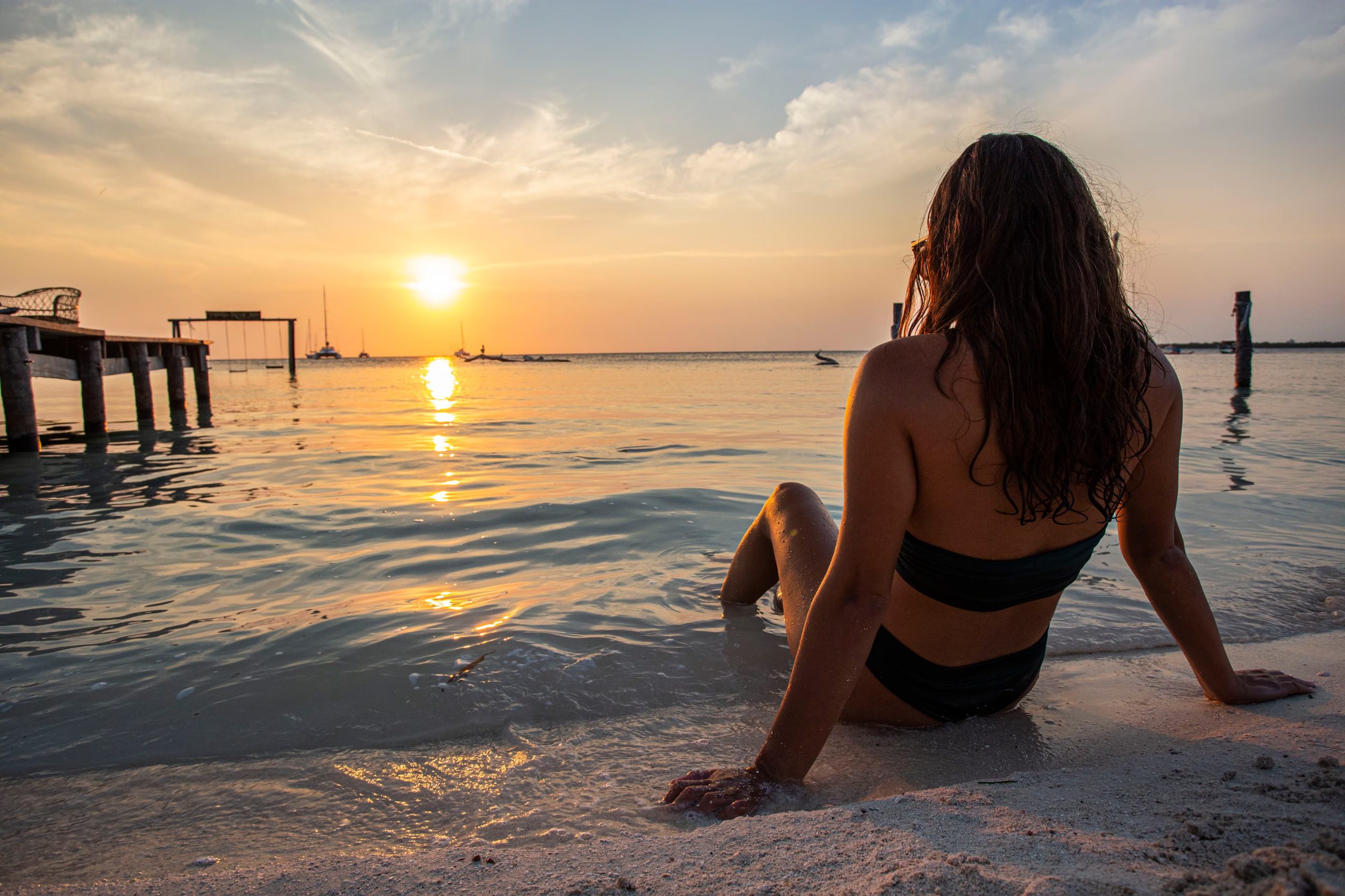 Una mujer de espaldas se sienta en la arena de la playa con los pies en el agua, mirando el sol que se oculta sobre el mar al atardecer. (The Belize Tourism Board (BTB), órgano del Ministerio de Turismo y Relaciones con la Diáspora de Belice.)