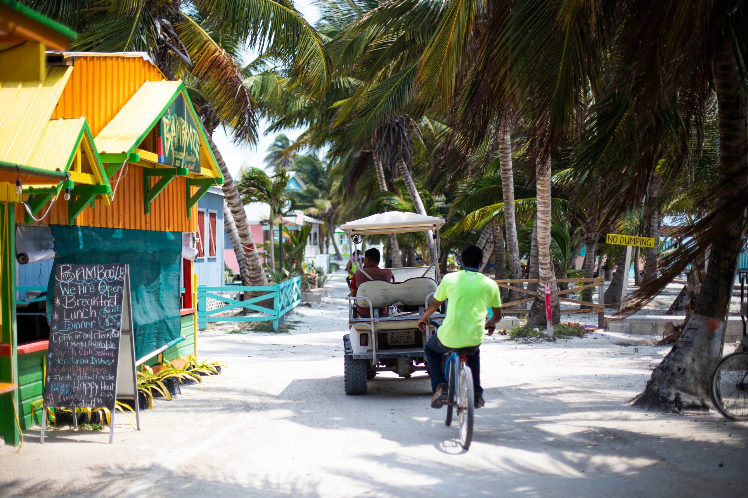 Una calle de arena en un entorno tropical con palmeras muestra a personas que se movilizan en bicicleta y en un carro de golf junto a edificaciones coloridas. (The Belize Tourism Board (BTB), órgano del Ministerio de Turismo y Relaciones con la Diáspora de Belice.)